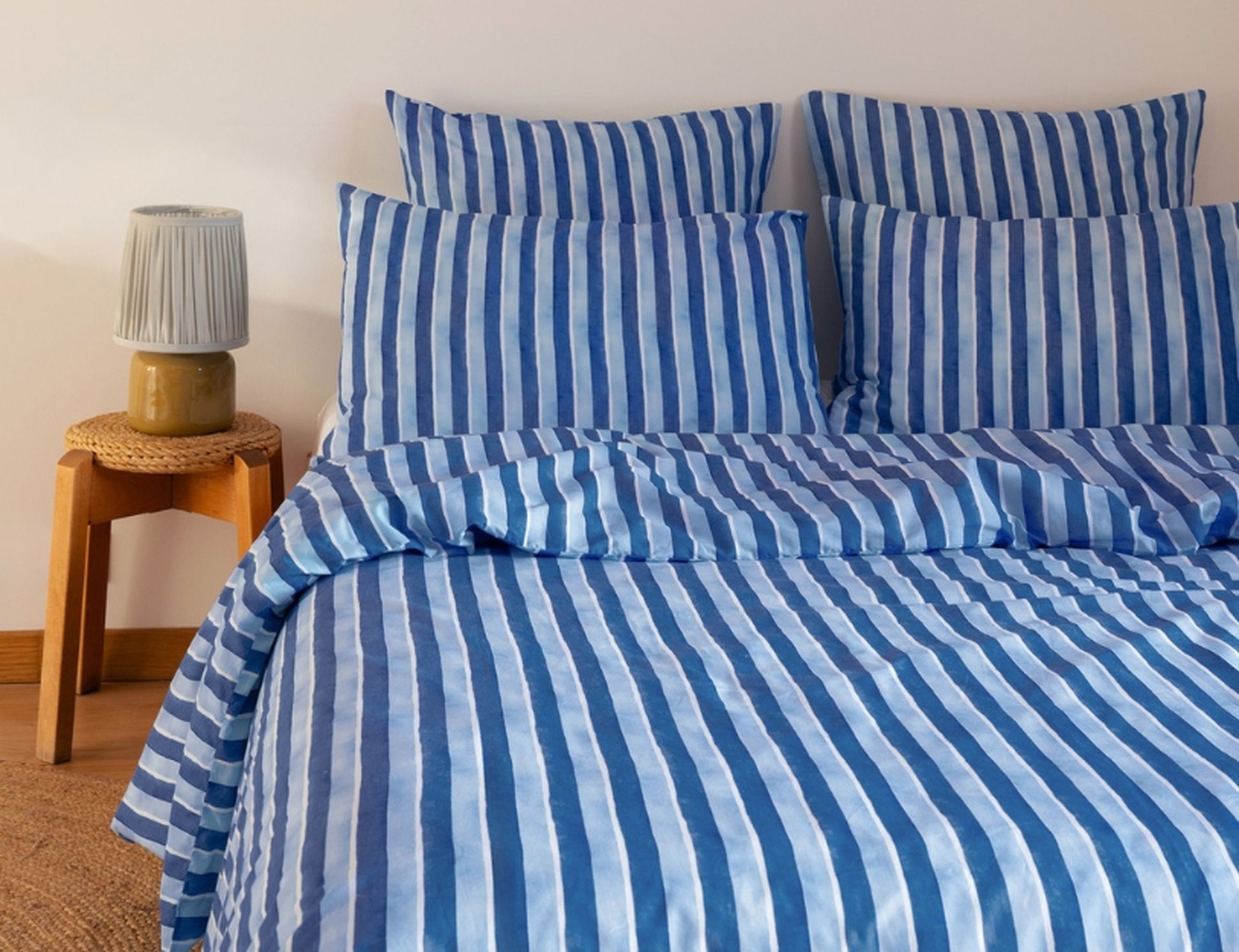 Un lit avec des draps et des oreillers à rayures bleues et blanches, à côté d'une table de chevet en bois sur laquelle se trouve une lampe.