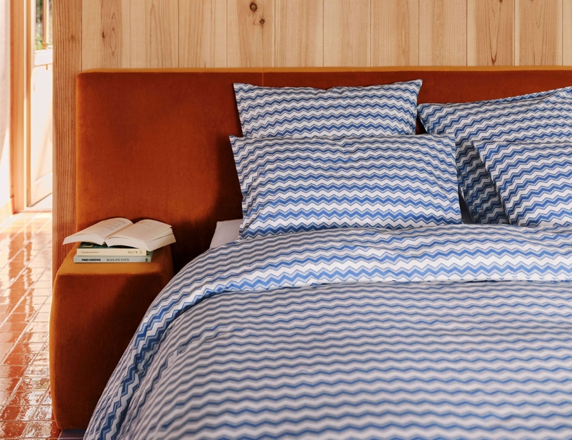 Un lit avec une parure de lit à motifs en zigzag bleus et blancs, une tête de lit orange et une table de chevet sur laquelle sont posés des livres ouverts, le tout devant un mur en bois.
