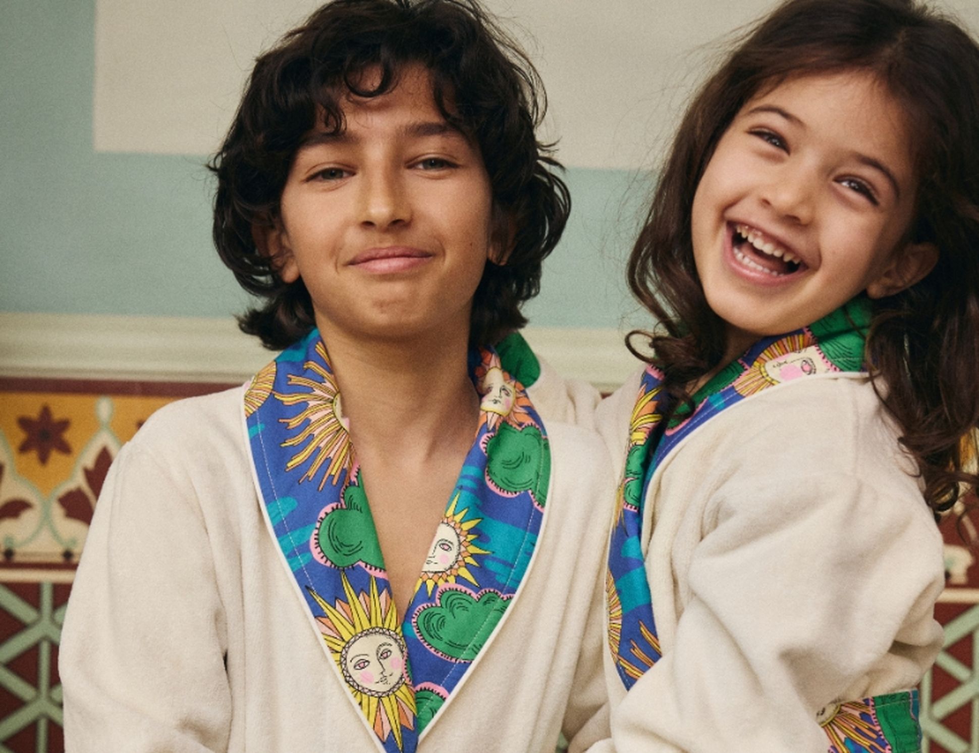 Deux enfants souriants, vêtus de peignoirs ornés de motifs colorés représentant des soleils et des nuages, se tiennent dans une pièce aux carreaux décoratifs.