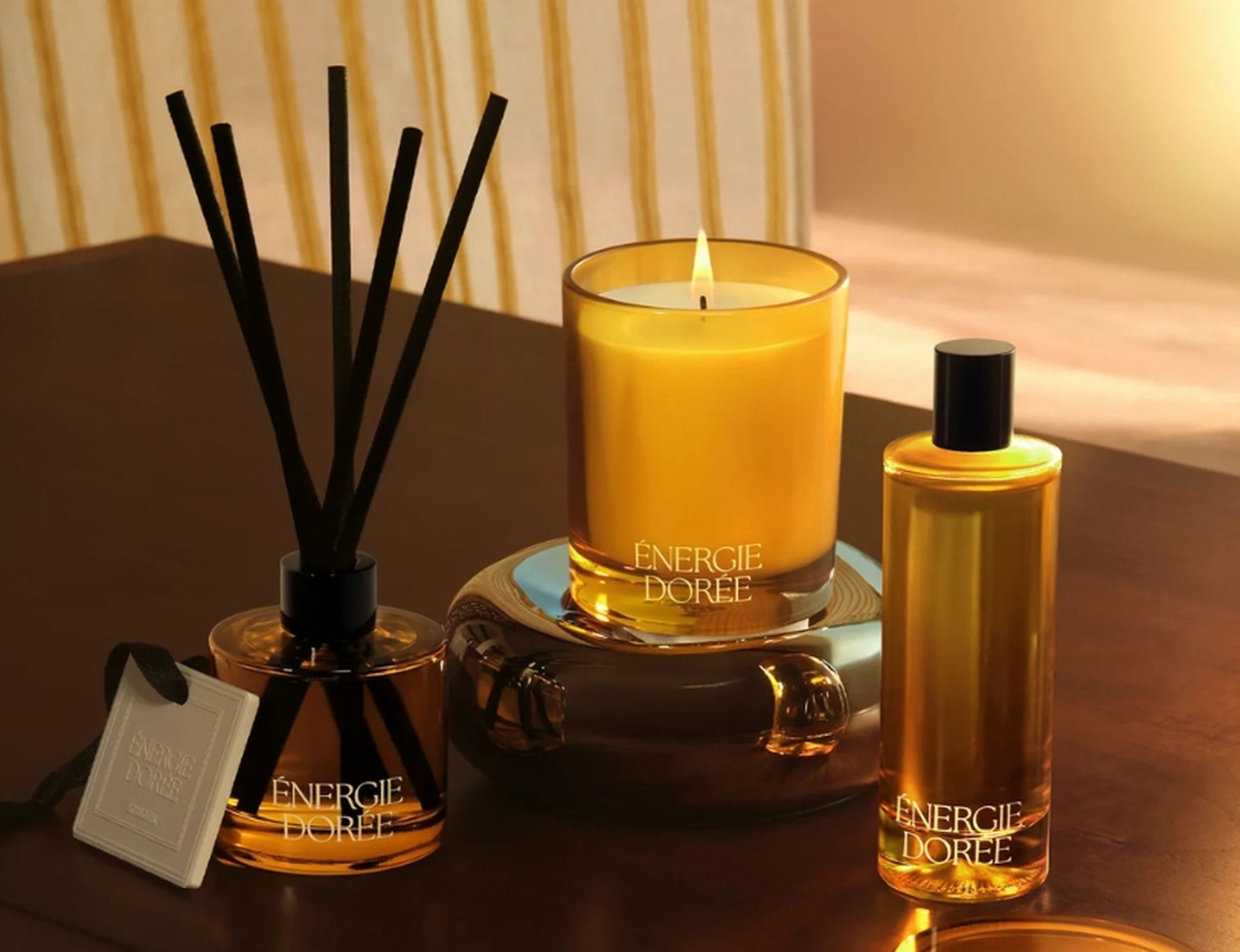 Une bougie dorée allumée, un diffuseur à bâtonnets et un flacon de lotion assorti portant l'inscription « Énergie Dorée » posés sur une table en bois, créant une ambiance chaleureuse et feutrée.