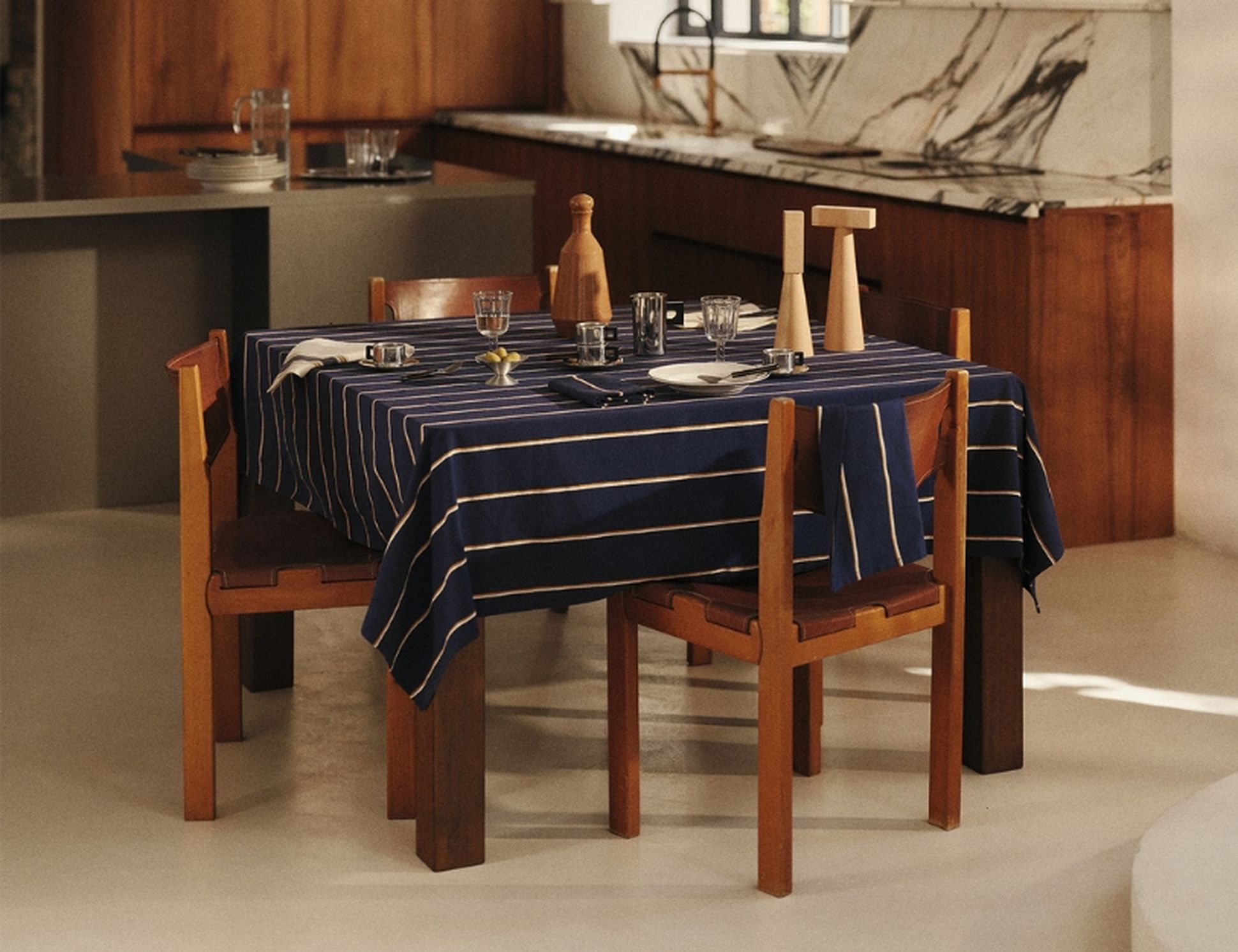 Une table à manger en bois recouverte d'une nappe à rayures bleues, quatre chaises et de la vaisselle, dans une cuisine moderne dotée de plans de travail en marbre.