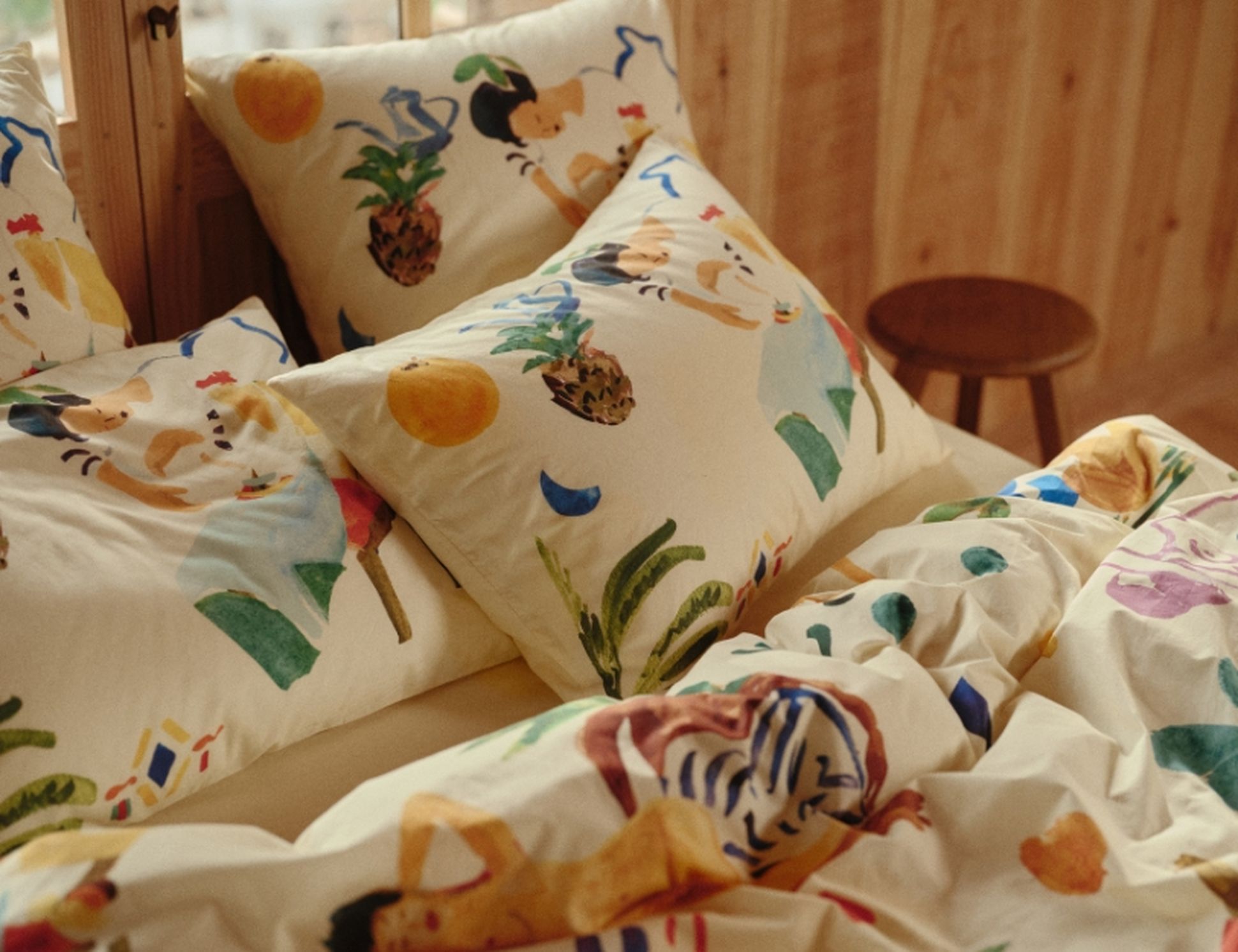 Coussins et linge de lit colorés, ornés de motifs ludiques et illustrés de fruits et de personnages, disposés sur un lit en bois chaleureux.