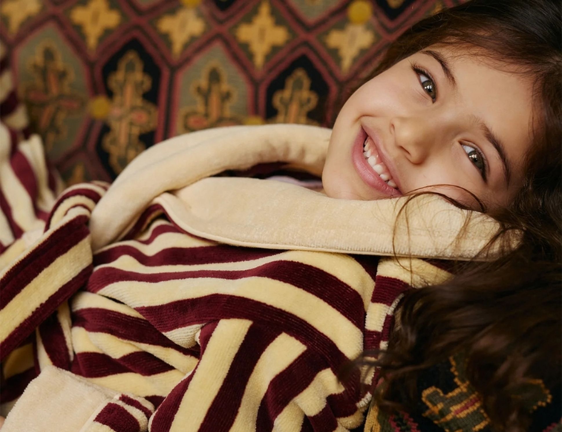 Un enfant souriant, vêtu d'un peignoir douillet à rayures bordeaux et beiges, allongé sur des coussins à motifs, avec de longs cheveux bruns et une expression joyeuse.