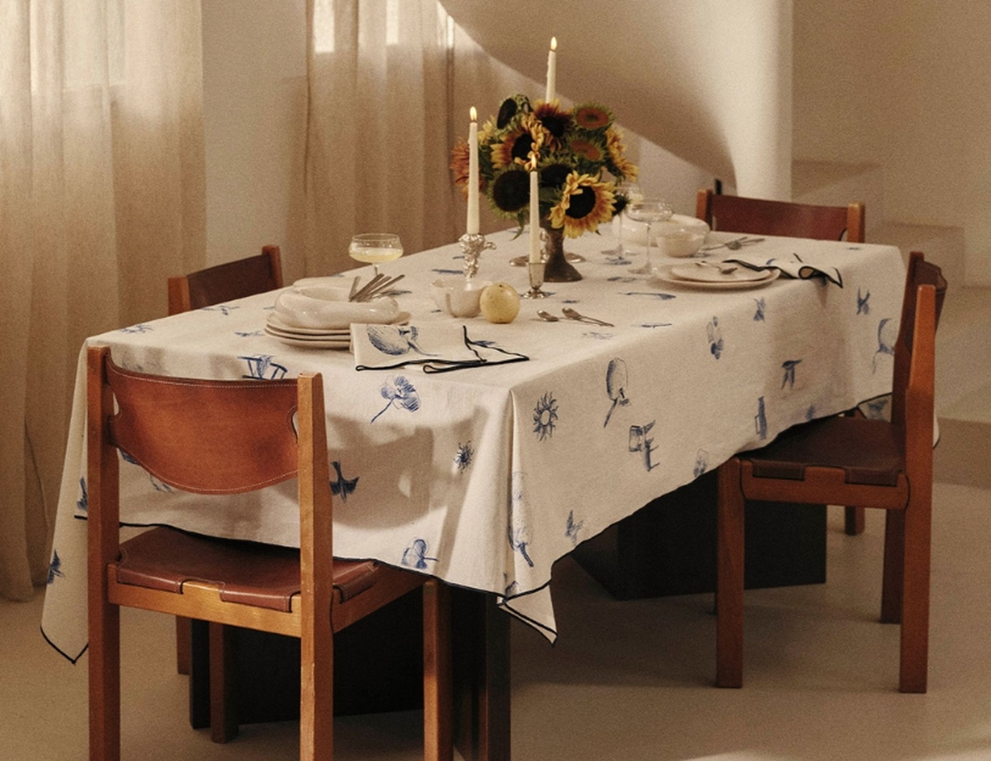 Une élégante table à manger recouverte d'une nappe blanche ornée de motifs bleus, dressée avec des bougies, des assiettes, des couverts et un bouquet éclatant de tournesols.