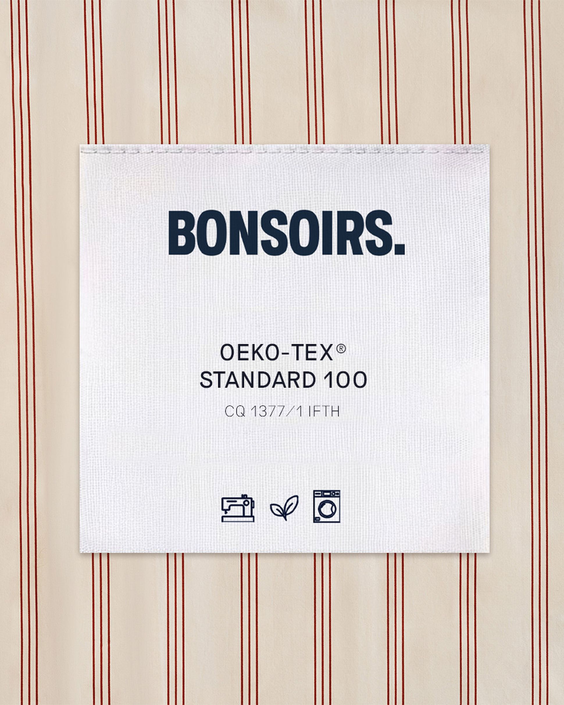Une étiquette portant la mention « BONSOIRS. OEKO-TEX STANDARD 100 » est cousue sur un tissu à rayures verticales rouges. Des icônes indiquent les consignes d'entretien et les normes écologiques.