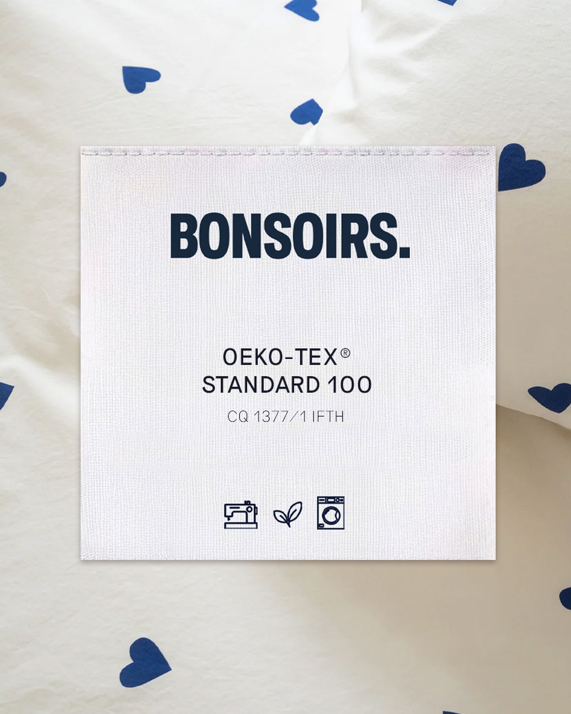 Une étiquette en tissu portant la mention « BONSOIRS. OEKO-TEX® STANDARD 100 » sur du linge de lit blanc orné de petits cœurs bleus. Des icônes indiquant les consignes de lavage figurent en dessous.