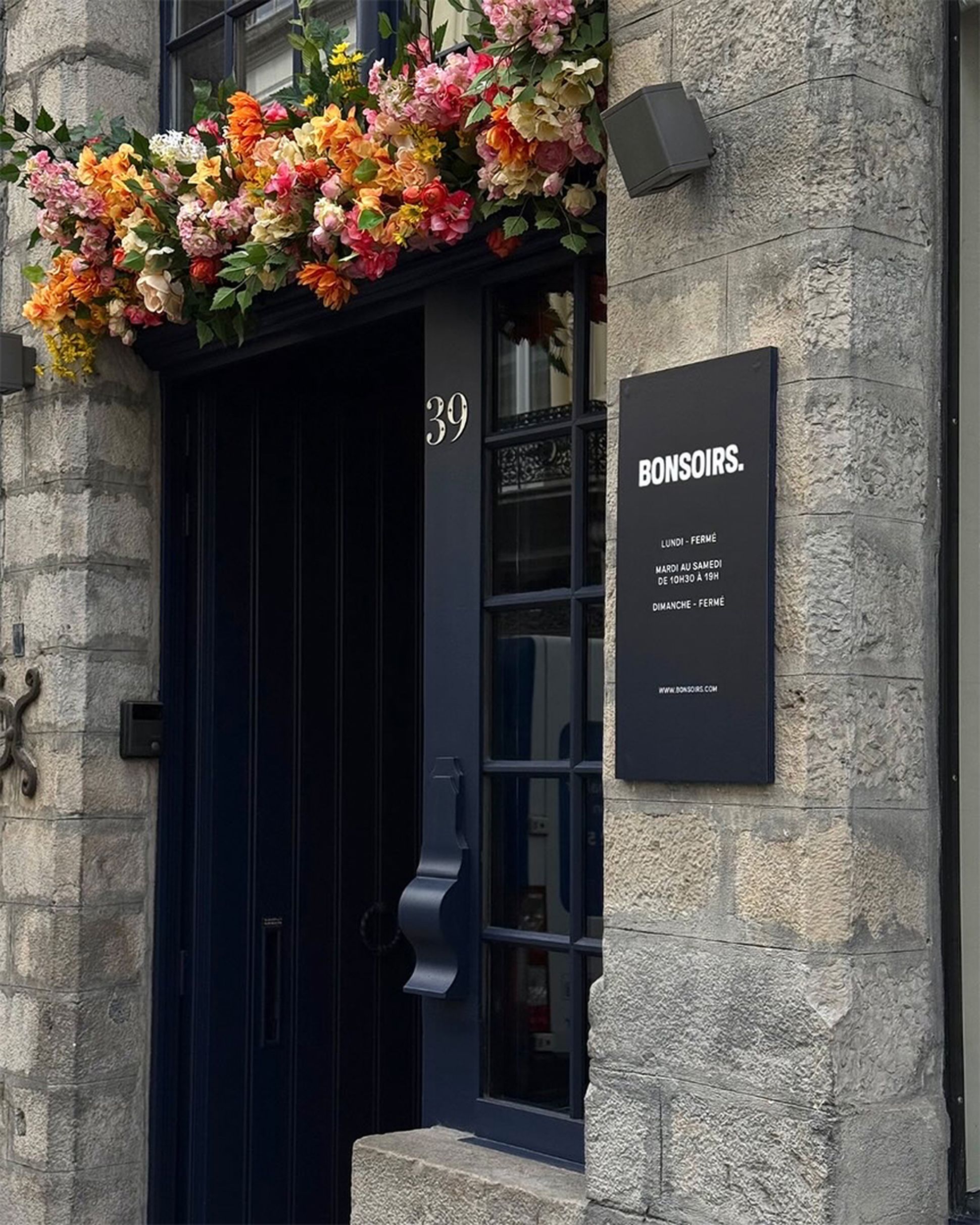 Porte bleu foncé surmontée d'une composition florale. Une enseigne indique « BONSOIRS » ainsi que les horaires d'ouverture. Façade en pierre sur laquelle on peut voir le numéro 39.