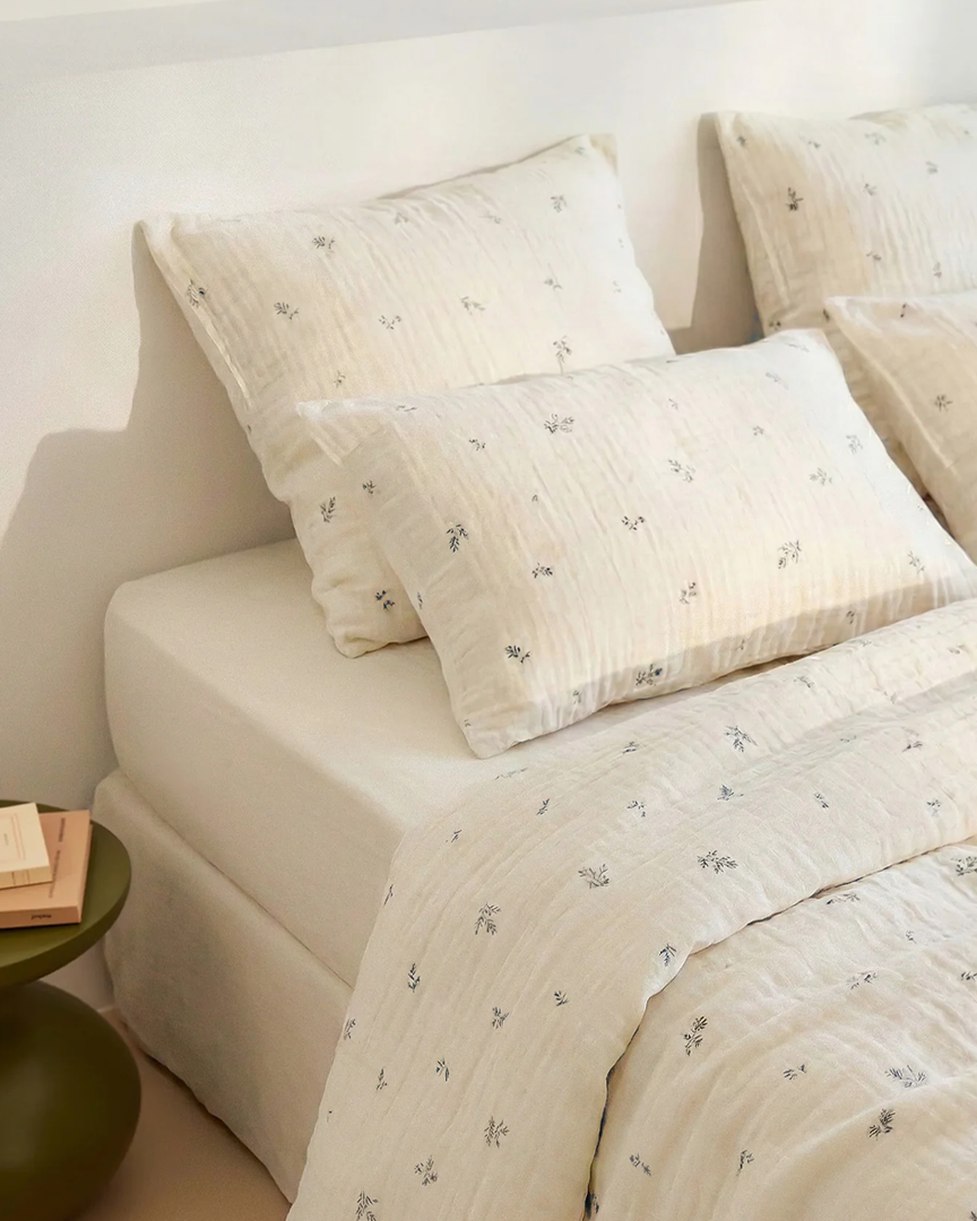 Un lit de couleur crème, avec des draps et des oreillers à motifs floraux, à côté d'une petite table de chevet verte sur laquelle sont posés des livres.