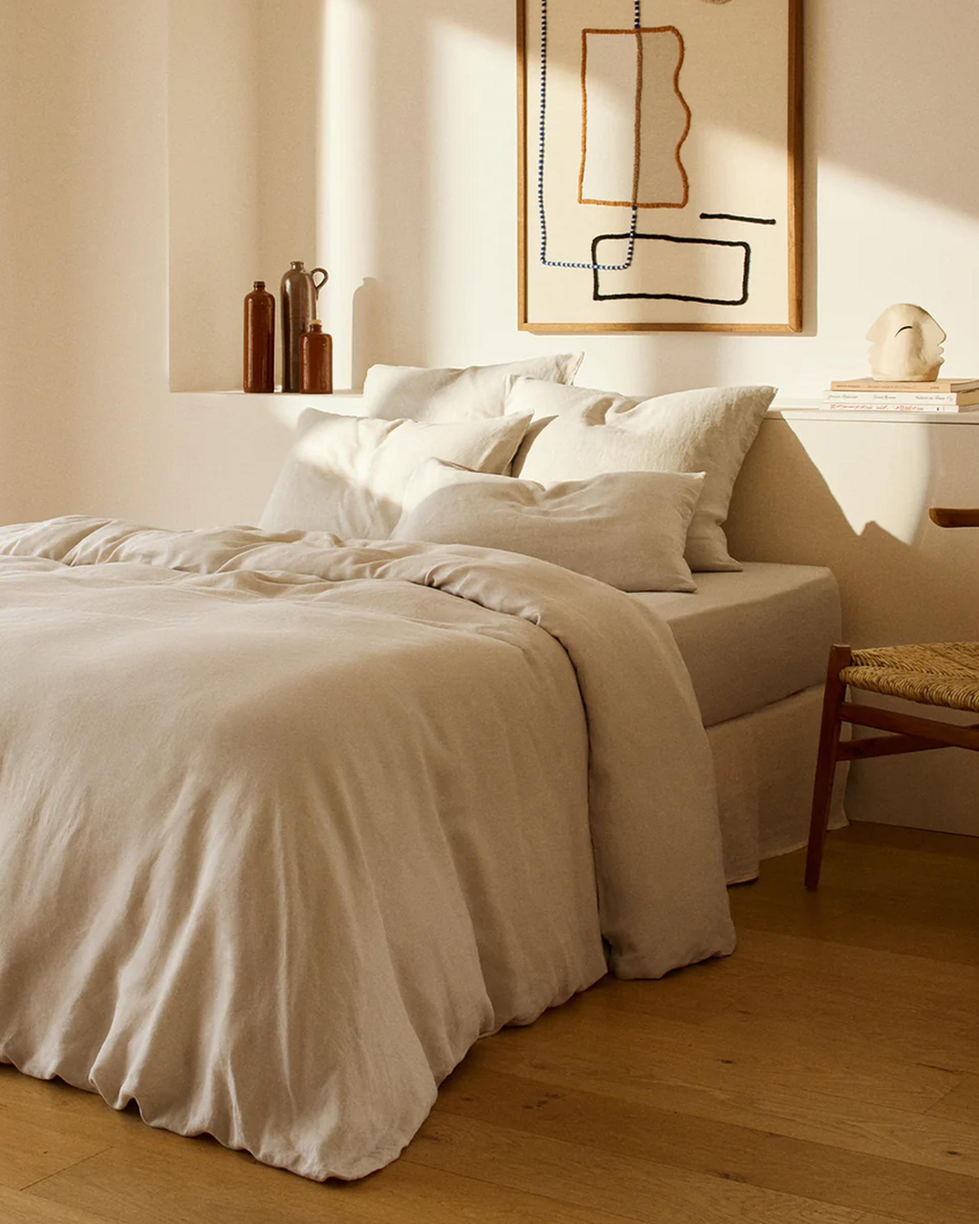 Une chambre minimaliste avec un lit beige, des oreillers blancs, des tableaux abstraits, des vases décoratifs, une petite sculpture et une chaise en bois posée sur un parquet.