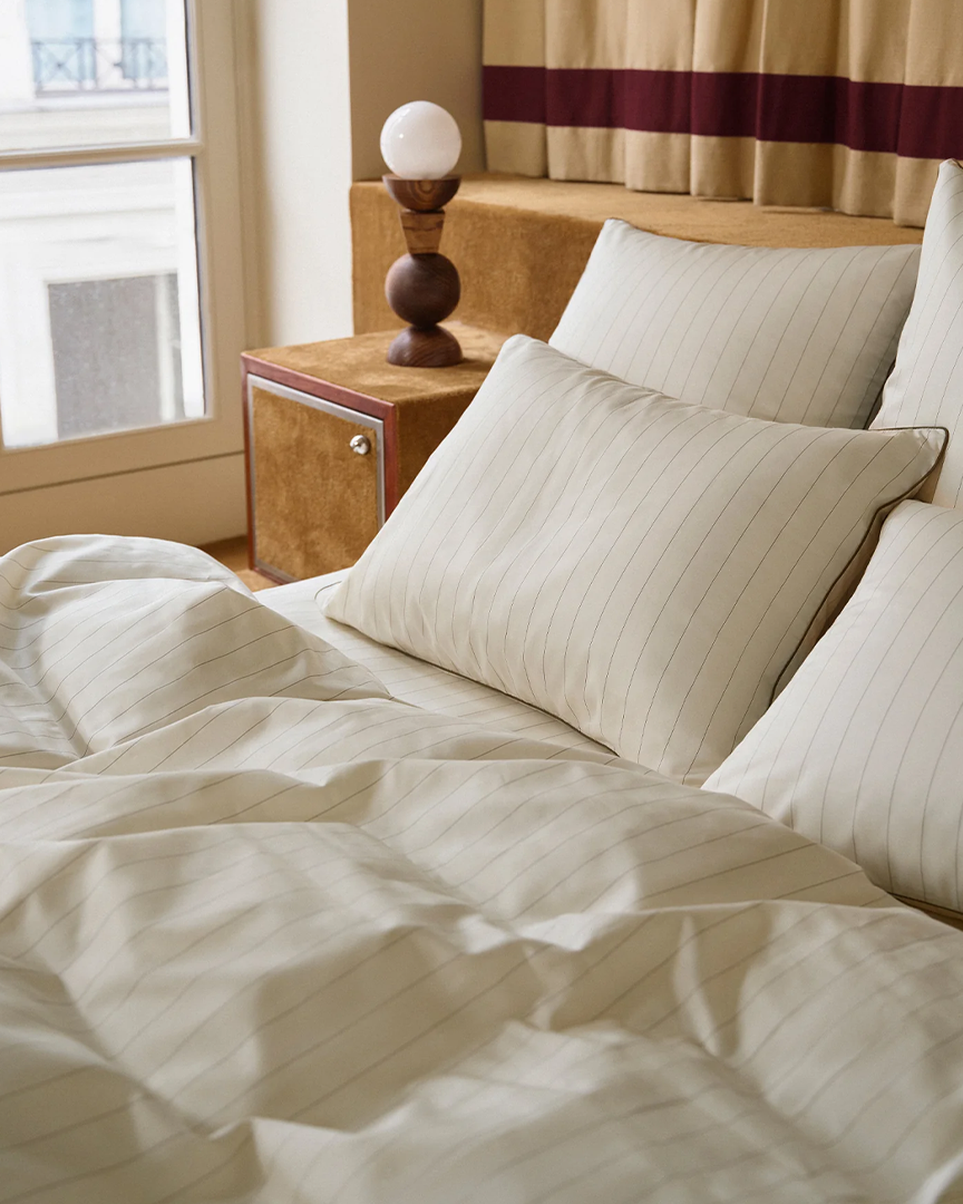 Un lit bien fait, avec des draps et des oreillers rayés de blanc, à côté d'une table de chevet en bois sur laquelle repose une lampe décorative. Une fenêtre en arrière-plan.