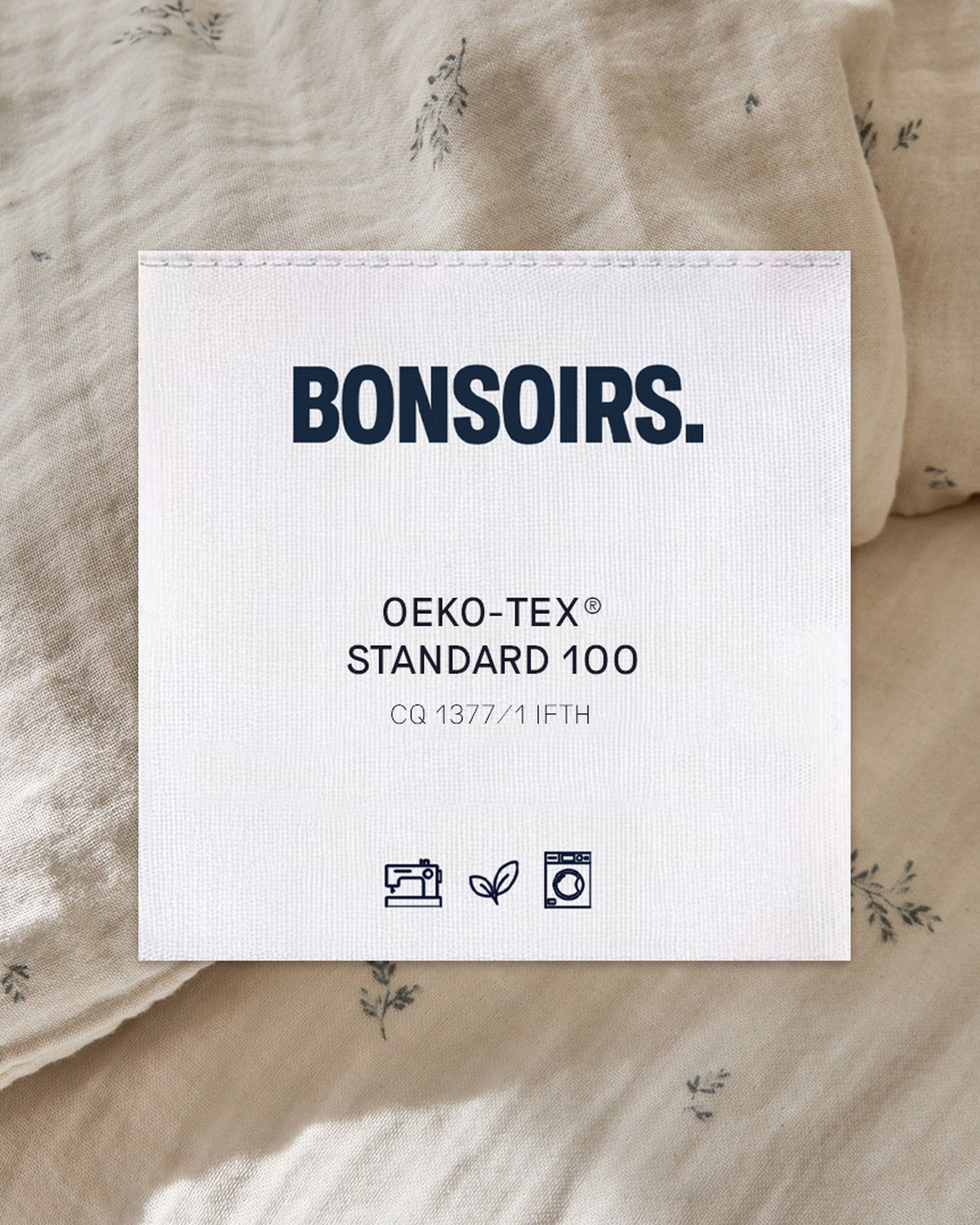 Une étiquette en tissu beige portant l'inscription « BONSOIRS » en caractères gras. Elle mentionne la certification « OEKO-TEX® STANDARD 100 » et comporte des symboles indiquant les consignes de lavage, de repassage et le respect de l'environnement.