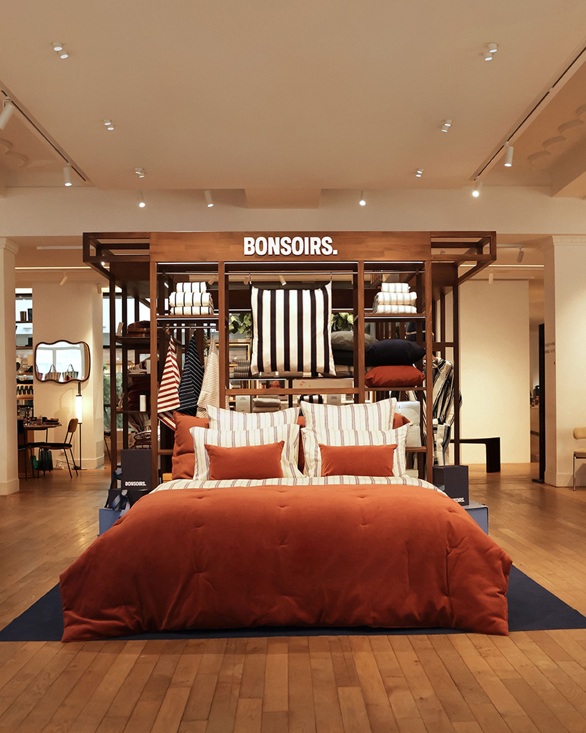 Dans un showroom chaleureux, on peut admirer un lit élégant, agrémenté d'une parure de lit couleur rouille et d'oreillers rayés, posé sur un parquet. Au-dessus, on aperçoit une enseigne « BONSOIRS. ».