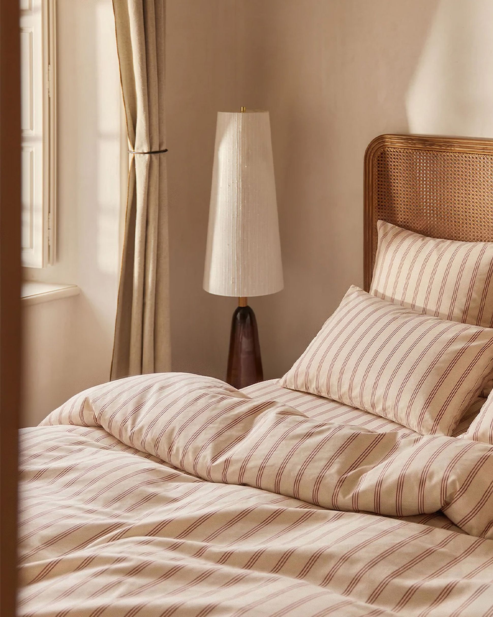 Une chambre douillette avec une parure de lit rayée, une tête de lit en rotin, un élégant lampadaire et des rideaux légers devant une fenêtre baignée de soleil.
