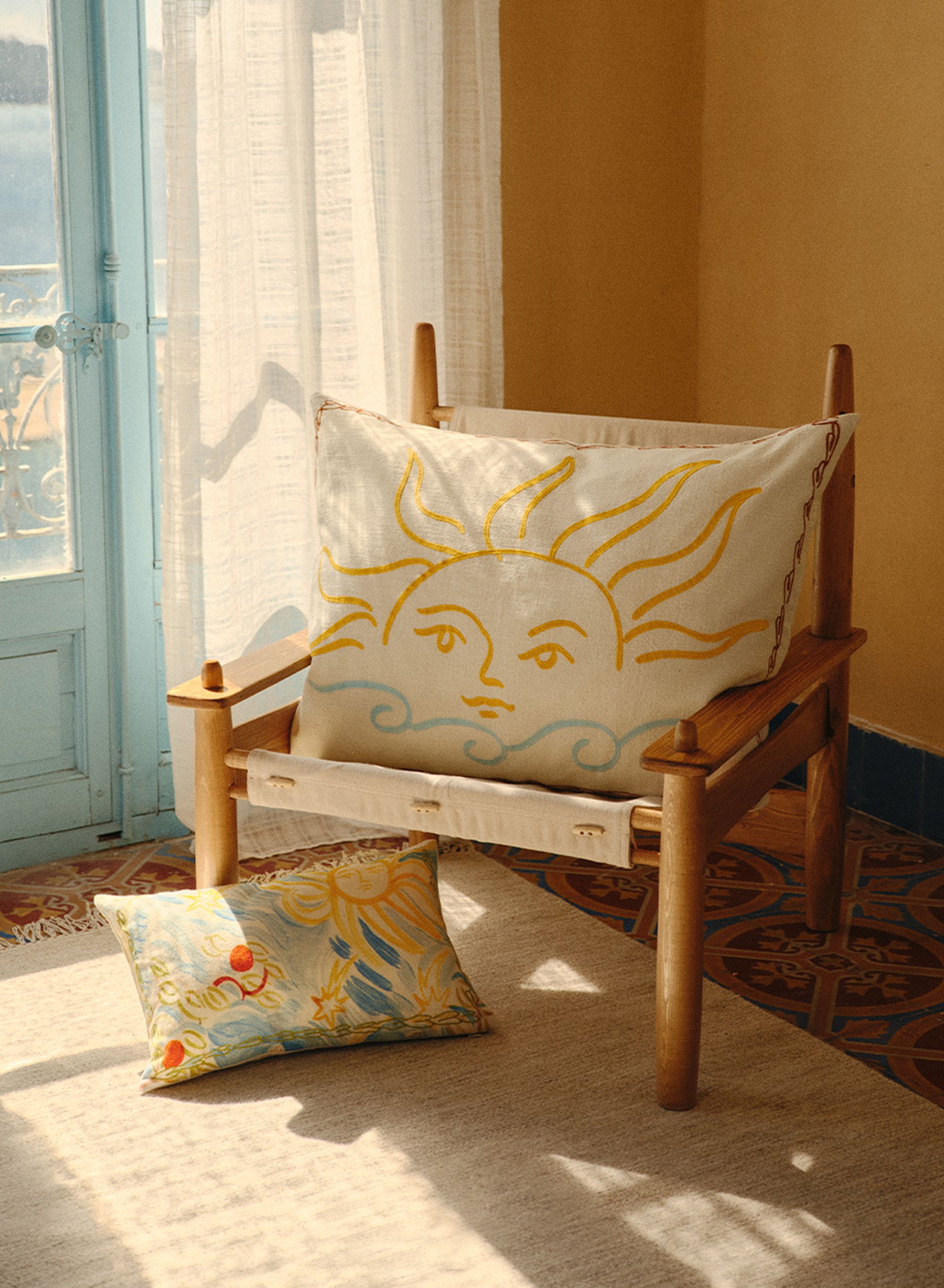 Chaise en bois avec un coussin sur le thème du soleil, baignée d'une lumière chaude près d'une fenêtre à encadrement bleu, posée sur un sol en carrelage à motifs.