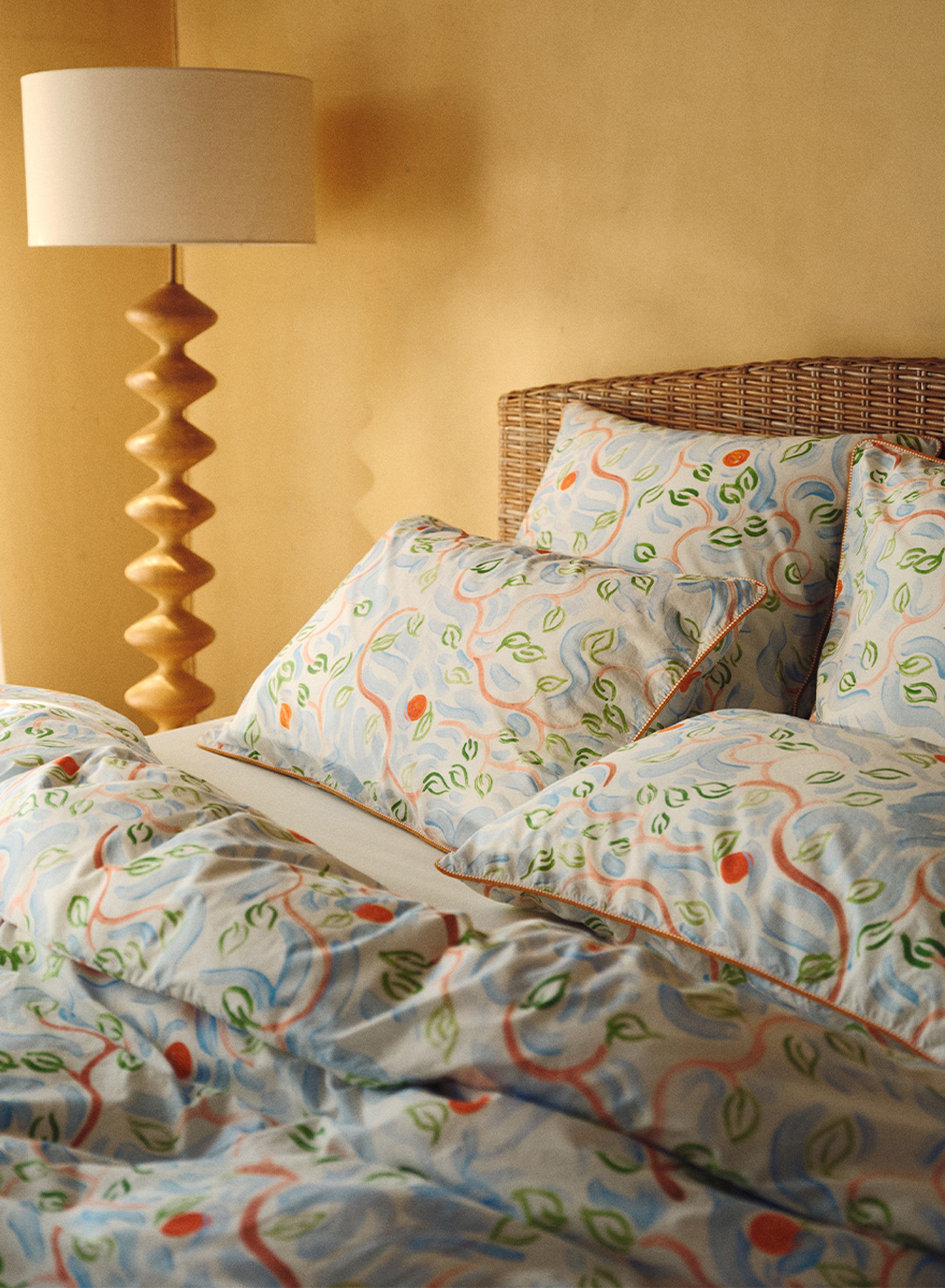 Un lit avec des draps et des oreillers aux motifs de feuilles colorés, une tête de lit en osier et une lampe de chevet en bois, devant un mur beige aux tons chauds.
