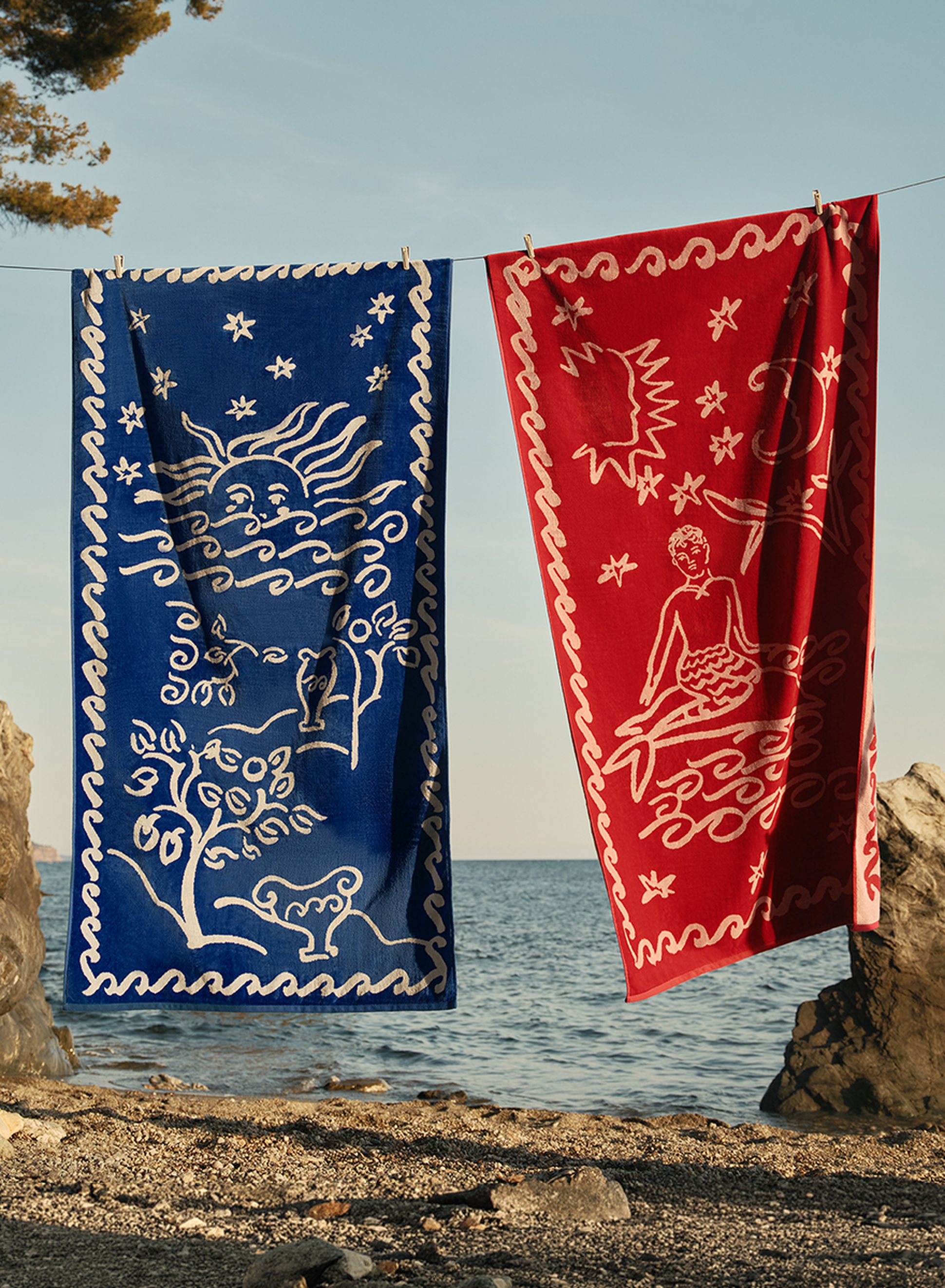 Deux serviettes aux couleurs vives, l'une bleue et l'autre rouge, ornées de motifs blancs inspirés du ciel et de l'océan, sont suspendues à une corde près de la mer.