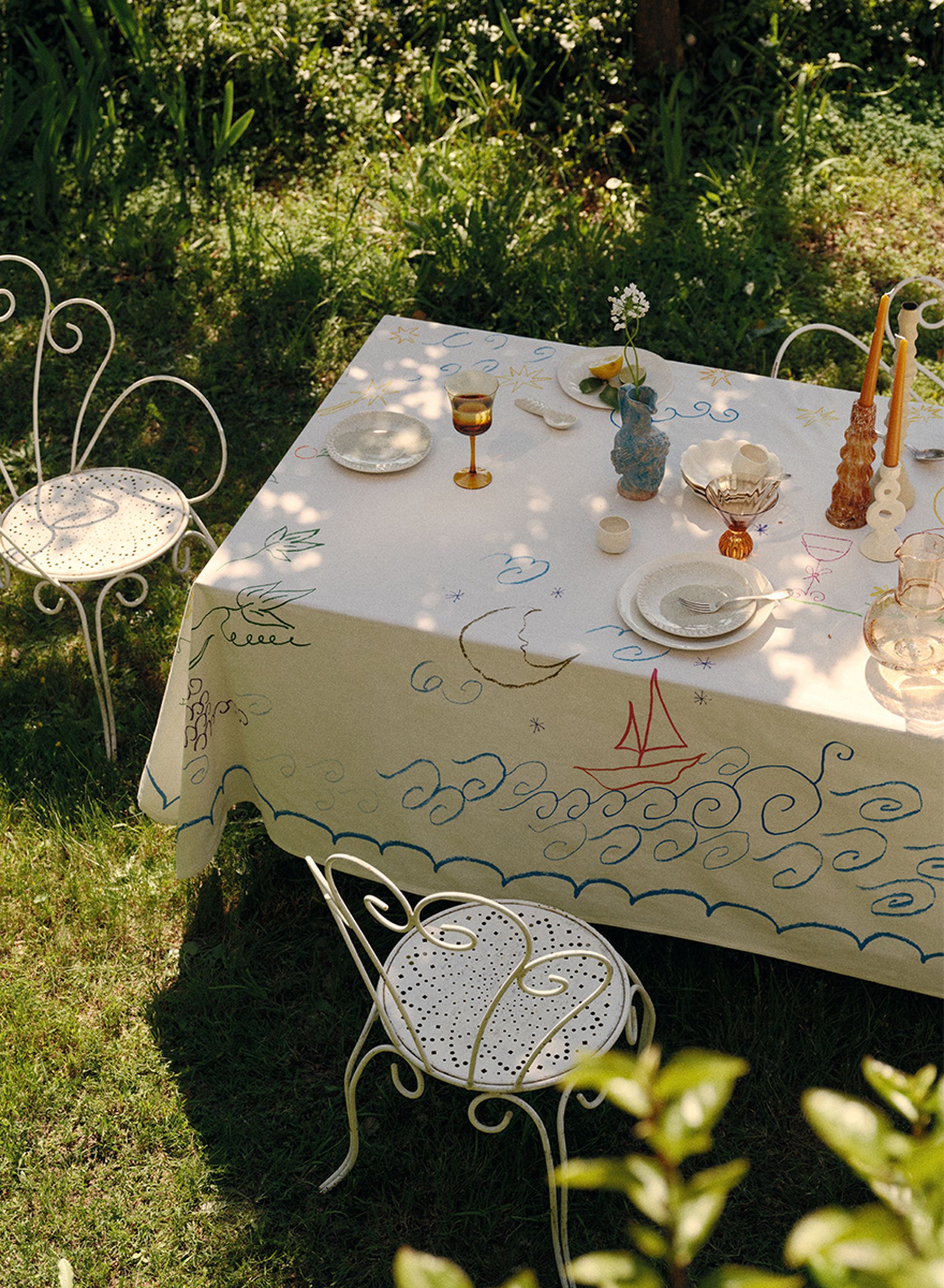 Une table d'extérieur recouverte d'une nappe sur le thème de la mer, dressée avec des verres et des couverts, entourée de chaises en fer forgé blanc sur une pelouse verte.