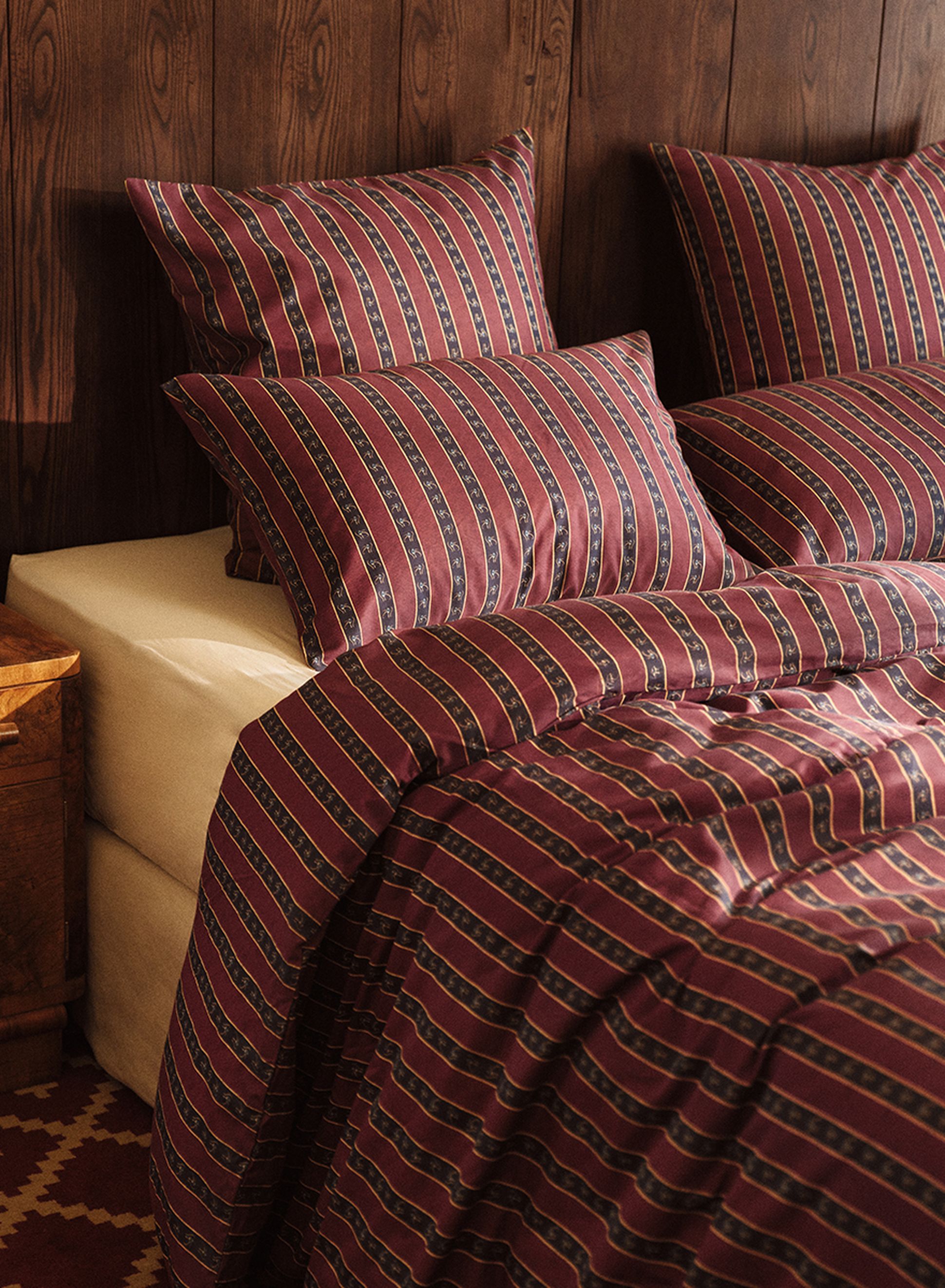 Un lit recouvert d'une parure de lit à rayures rouge foncé et bleu marine, comprenant des oreillers et une couette, adossé à un mur en bois, à côté d'une table de chevet en bois.