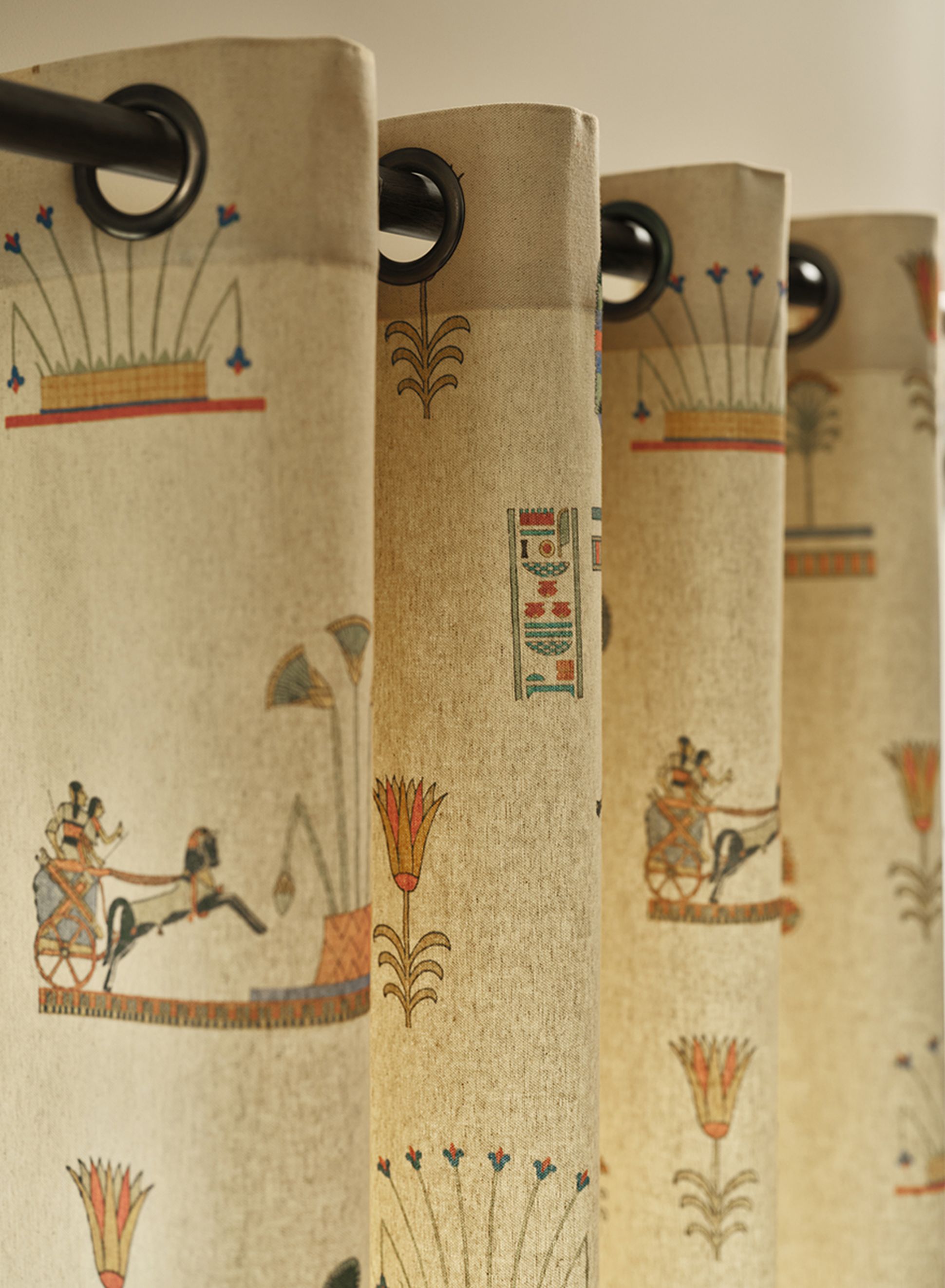 Des rideaux beiges ornés de motifs de style égyptien en rouge, bleu et vert, représentant des chars et des fleurs de lotus, suspendus à une tringle noire.