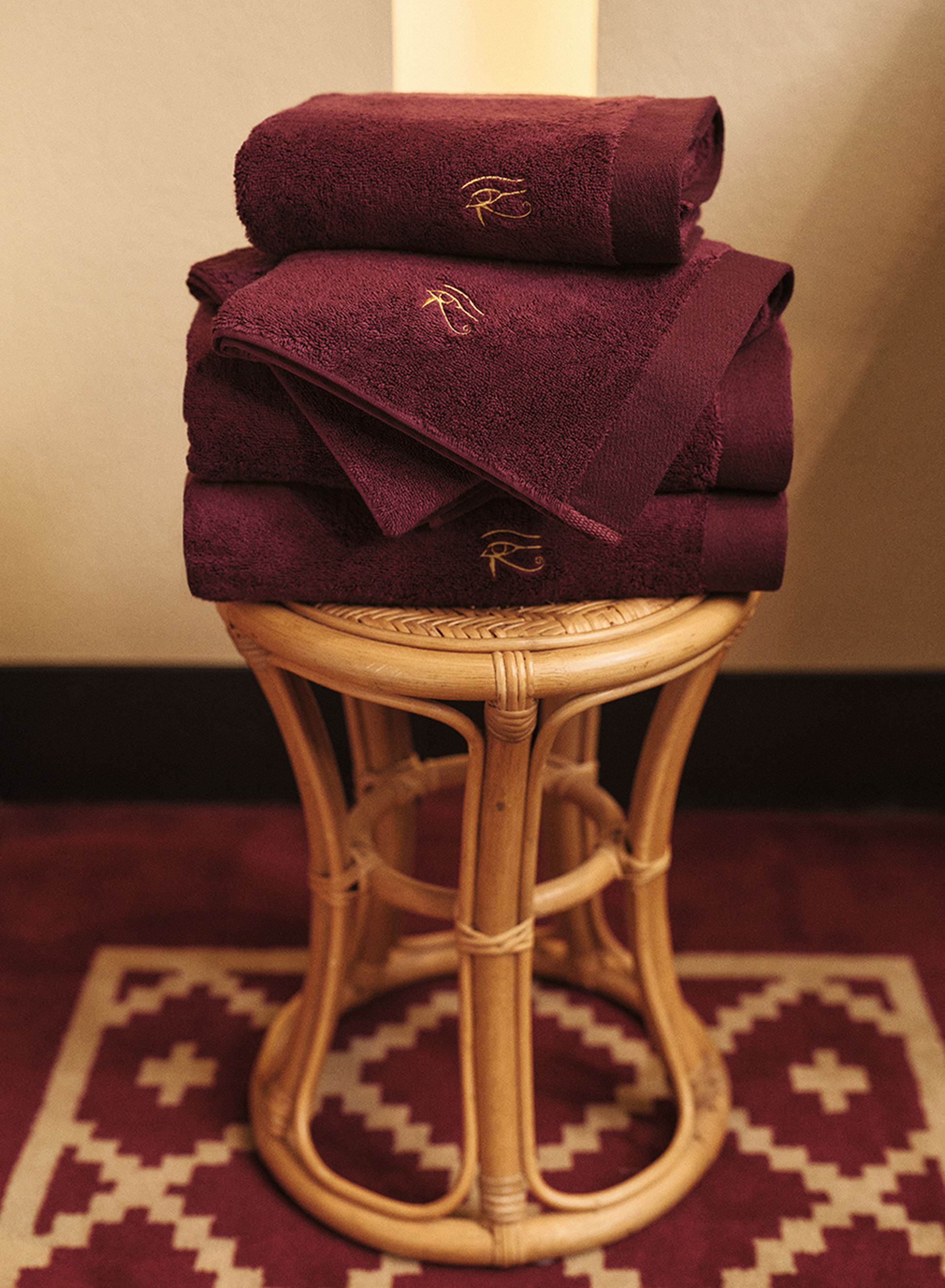 Une pile de serviettes bordeaux ornées de broderies dorées, posée sur un tabouret en osier, au milieu d'un tapis à motifs.