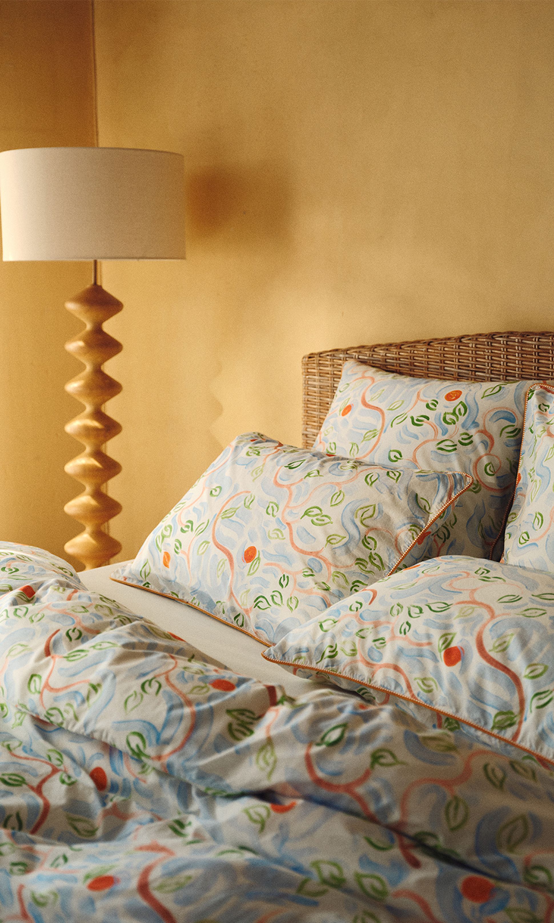 Lit orné d'oreillers à motifs et d'une couette aux volutes colorées et aux motifs de feuilles. Lampe en bois et tête de lit en osier devant un mur beige.