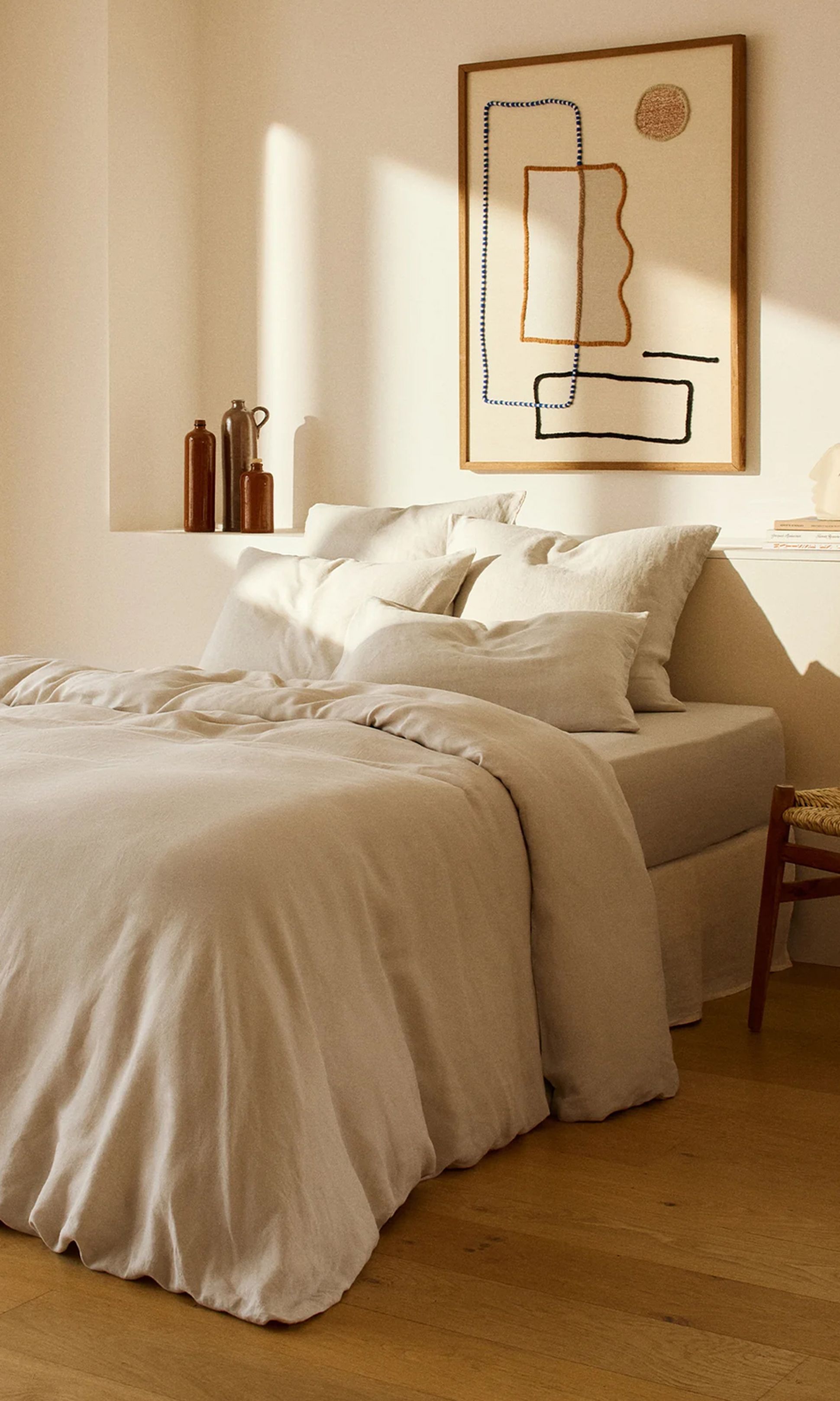 Chambre à coucher minimaliste de couleur beige, avec un lit soigneusement fait, des coussins douillets, des œuvres d'art abstraites accrochées au mur, un parquet et des bocaux décoratifs posés sur une étagère.