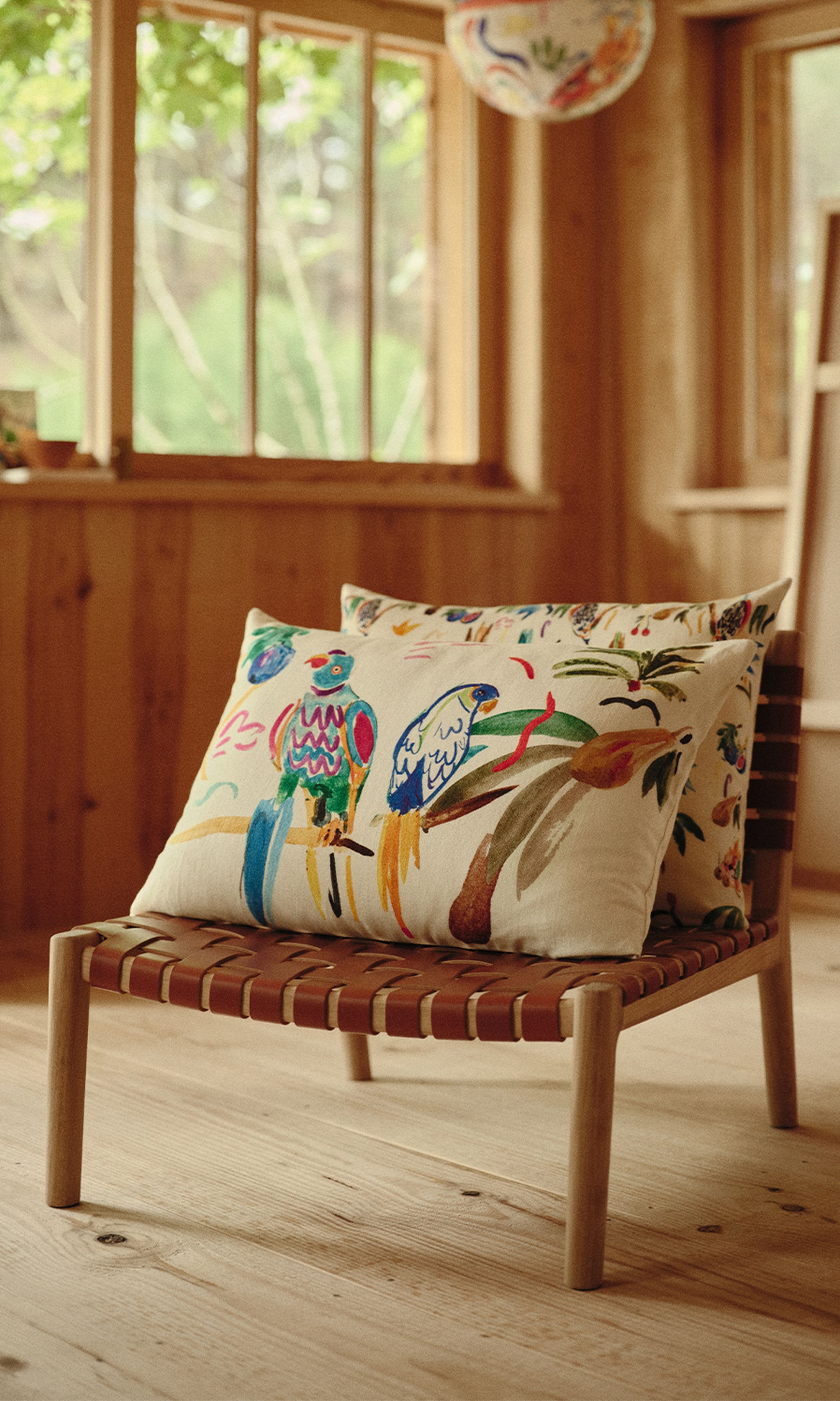 Une chaise en bois avec des coussins aux motifs colorés représentant des perroquets, dans une pièce baignée de soleil aux murs en bois et aux grandes fenêtres.