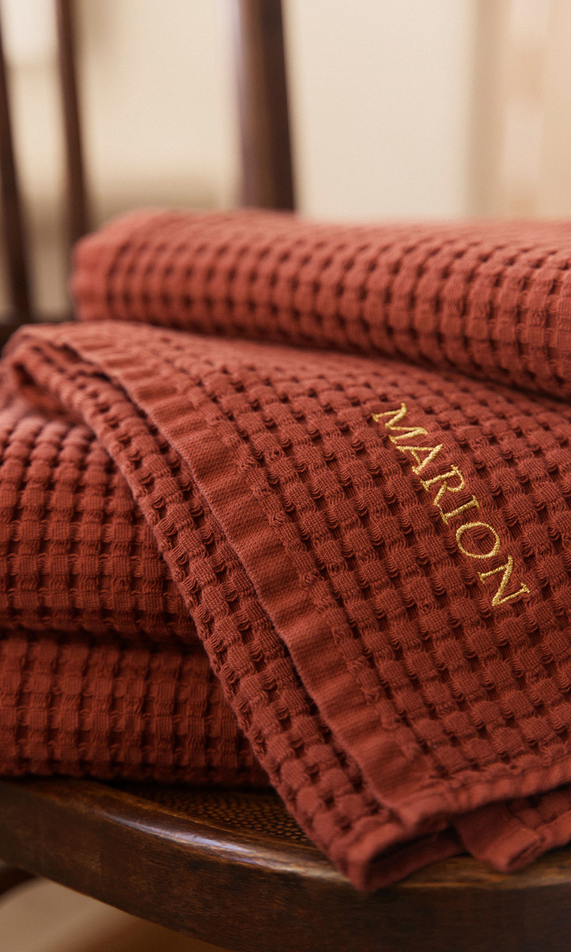 Pile de serviettes en maille gaufrée couleur rouille avec « MARION » brodé en doré, posée sur une chaise en bois.