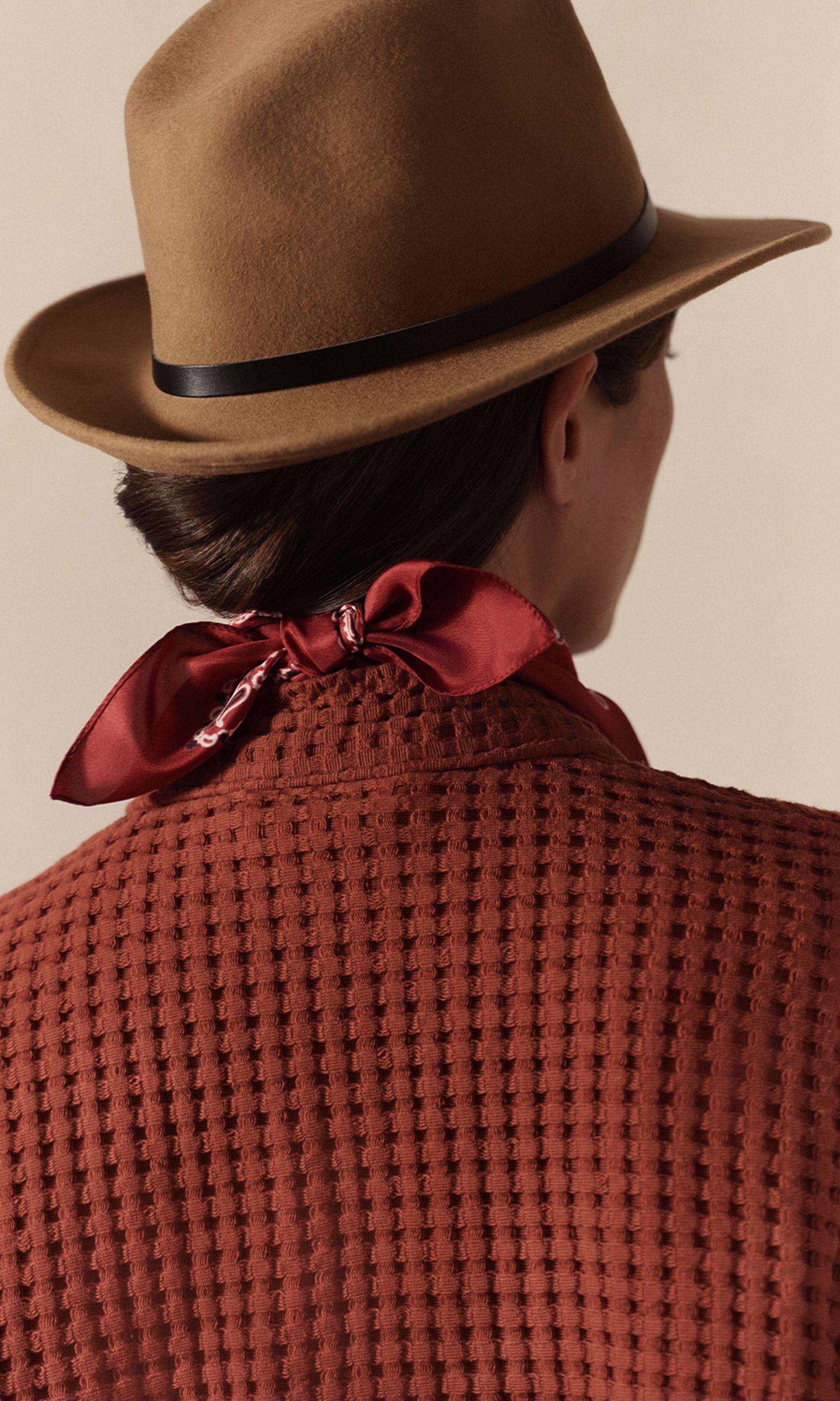 Personne portant un chapeau marron, un haut texturé couleur rouille et une écharpe rouge nouée dans le dos. Elle tourne le dos à la caméra.