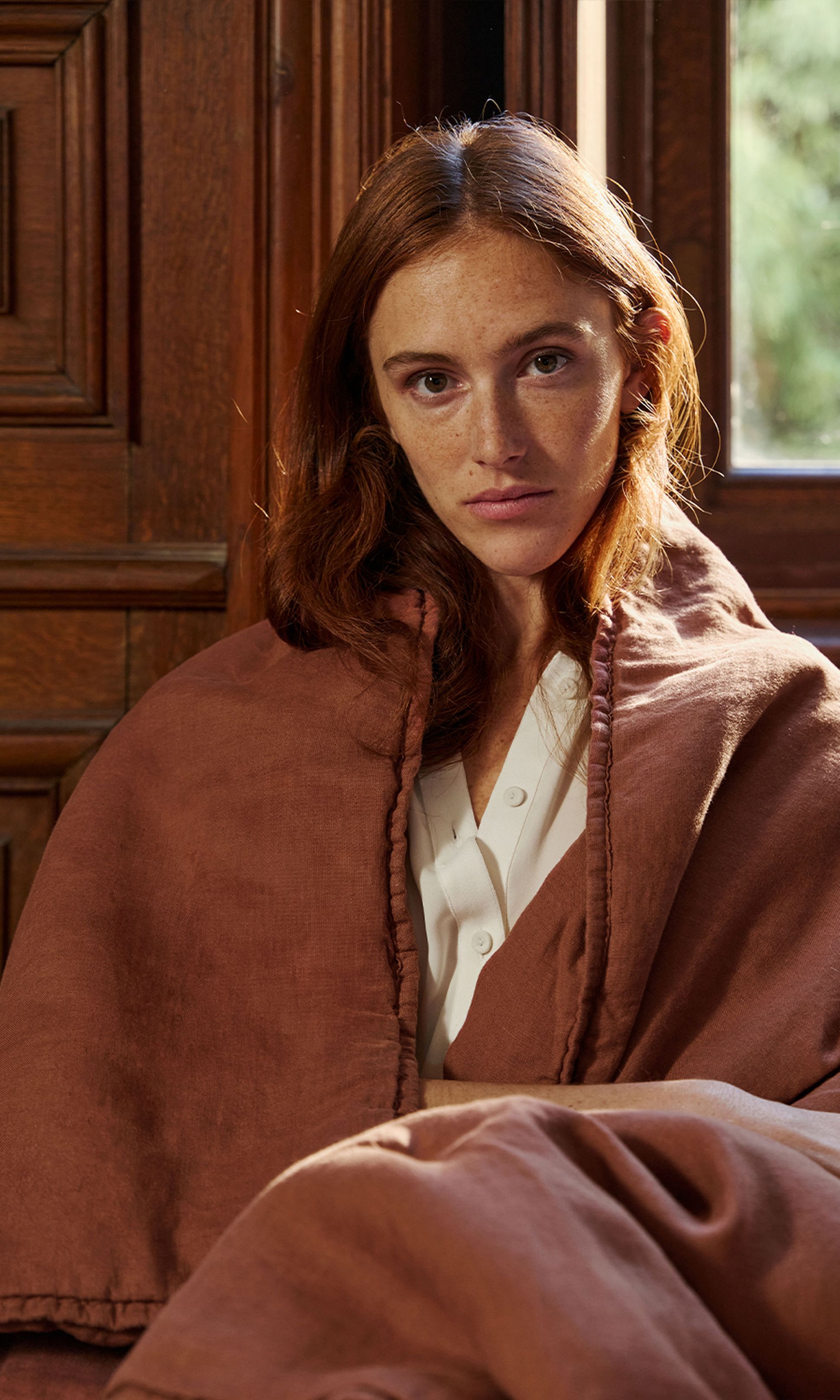 Une femme aux cheveux roux, enveloppée dans une couverture marron, assise à l'intérieur près d'un mur en bois, vêtue d'une chemise blanche, le regard rivé sur l'appareil photo.