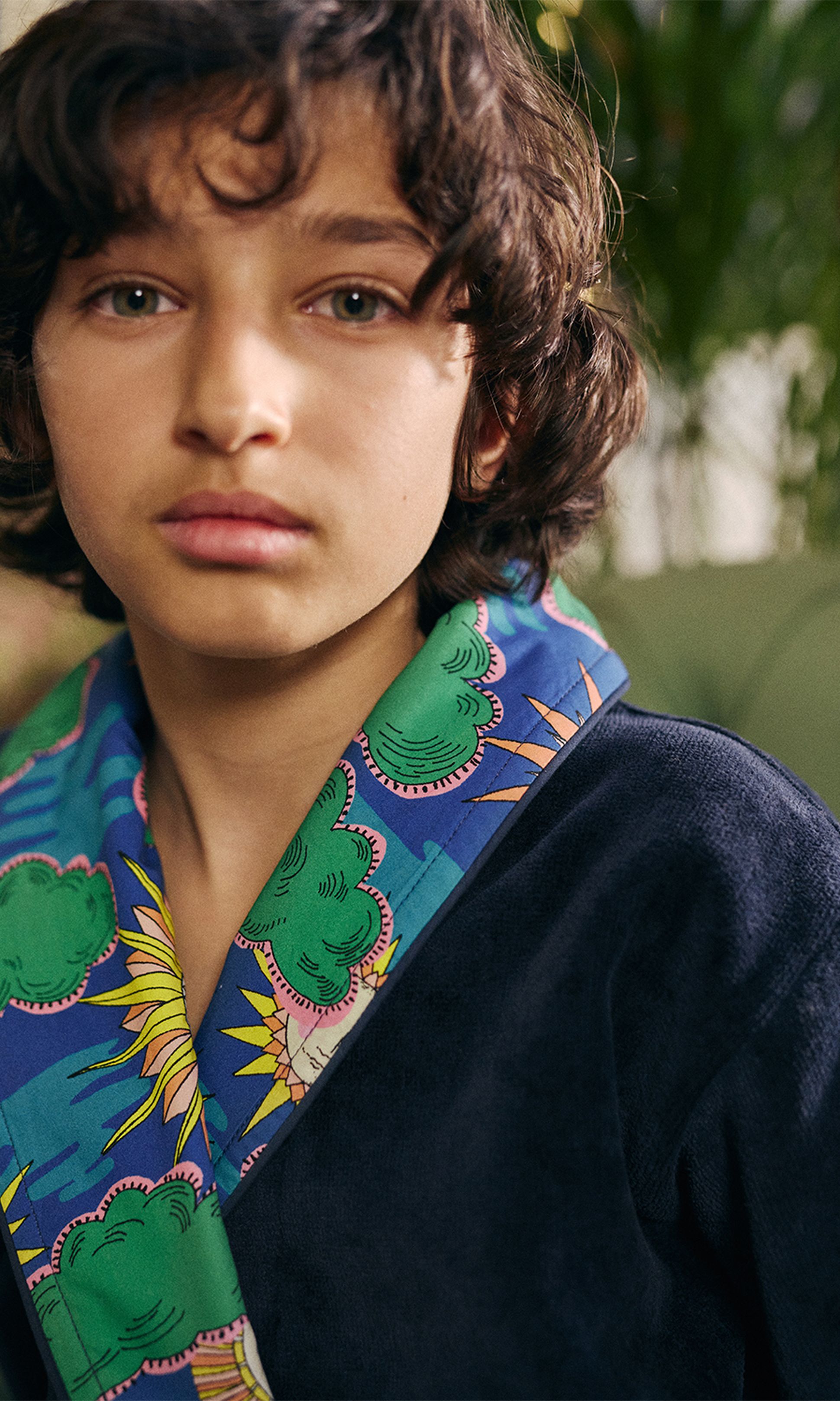 Gros plan sur une personne aux cheveux bouclés, vêtue d'un vêtement bleu orné d'un col à motifs colorés, sur fond vert flou.