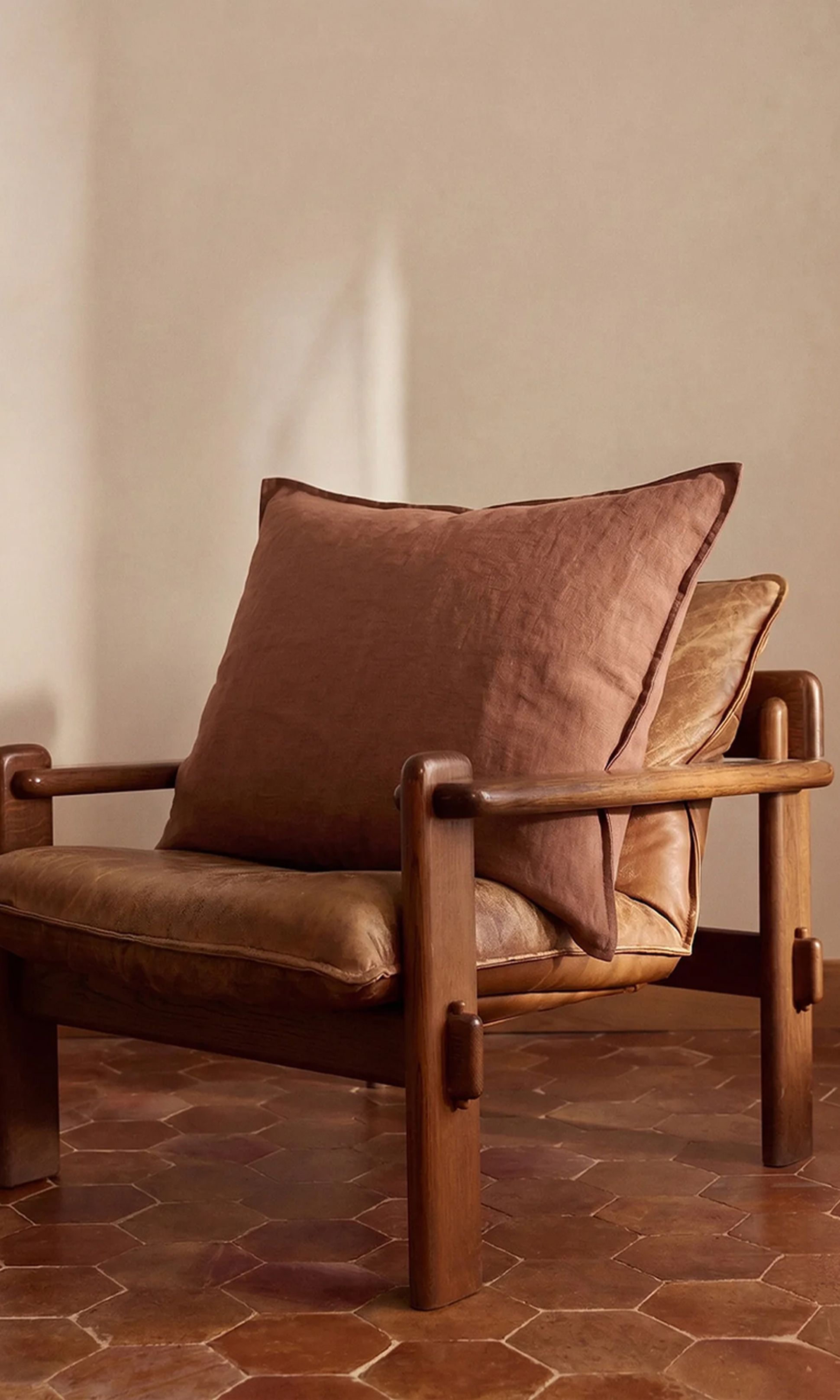 Un fauteuil vintage en bois, doté de coussins en cuir marron et d'un grand coussin marron assorti, posé devant un mur aux tons chauds et un sol en carreaux hexagonaux.