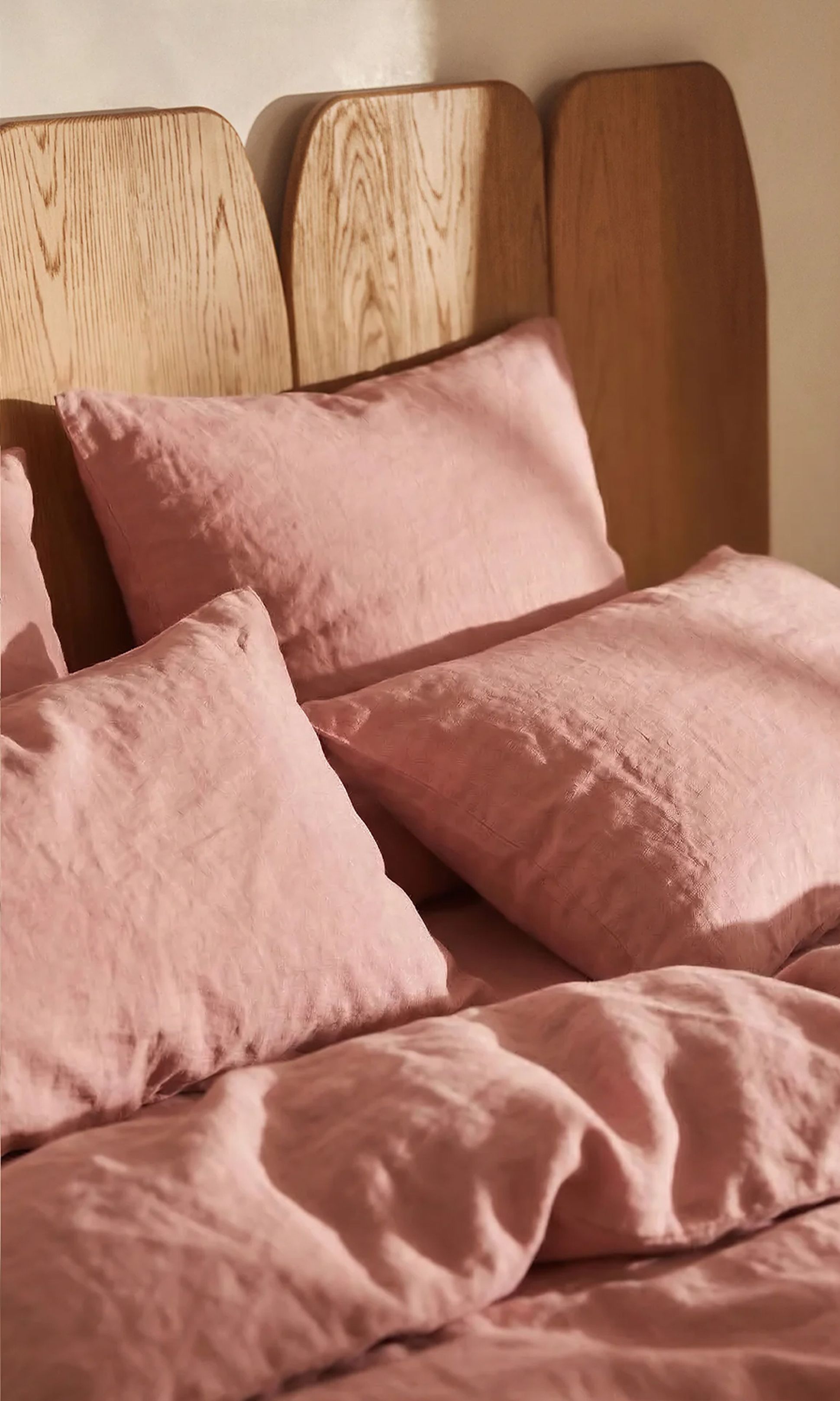 Un lit avec une tête de lit en bois, recouvert d'oreillers en lin rose pâle et d'une couette, baigné par la lumière chaude du soleil.
