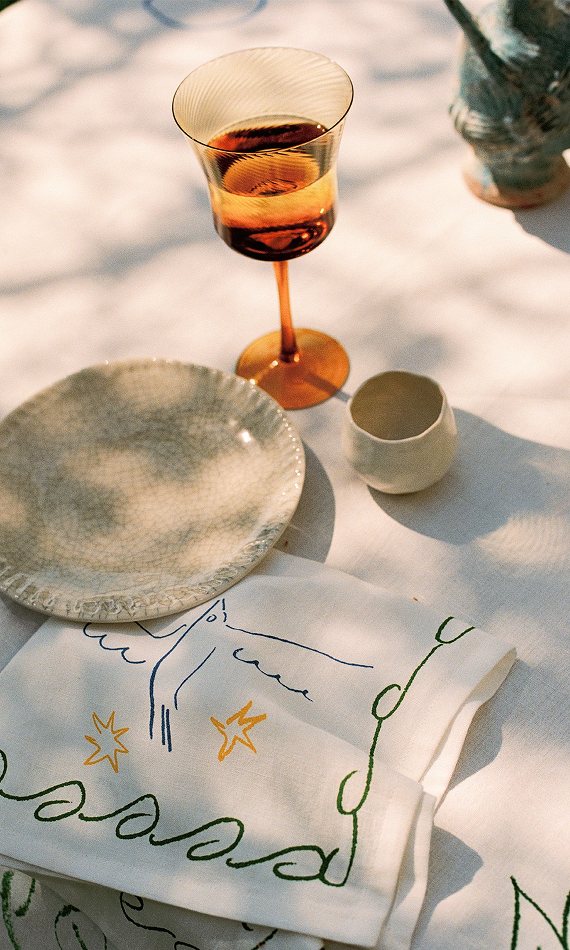 Une table élégamment dressée, avec une assiette texturée, un verre à vin ambré, une tasse en céramique et une serviette artistique posés sur une nappe baignée de soleil.