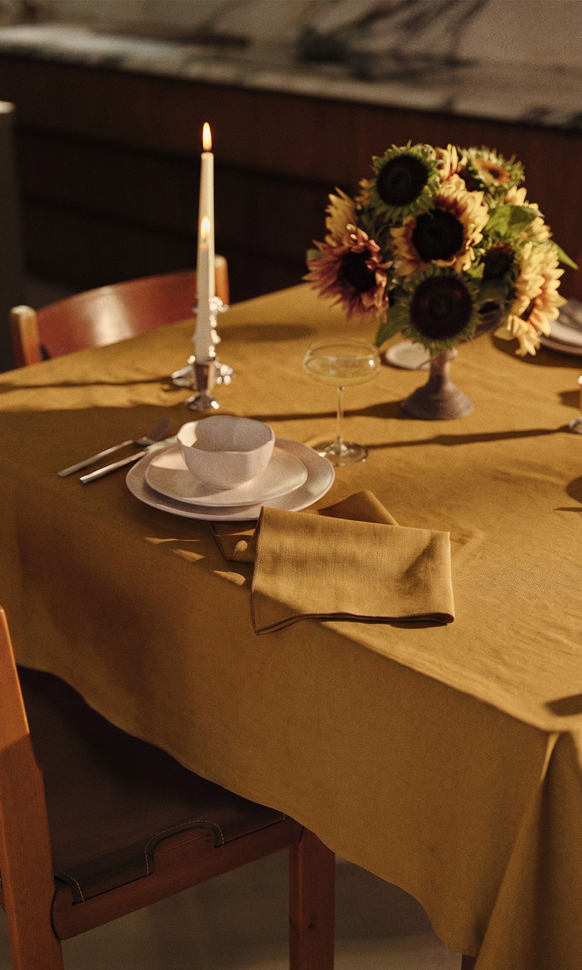 Une table à manger baignée d'une lumière chaleureuse, recouverte d'une nappe couleur moutarde, dressée avec un bol et une assiette, des couverts, une bougie allumée, une serviette pliée et un vase rempli de tournesols.
