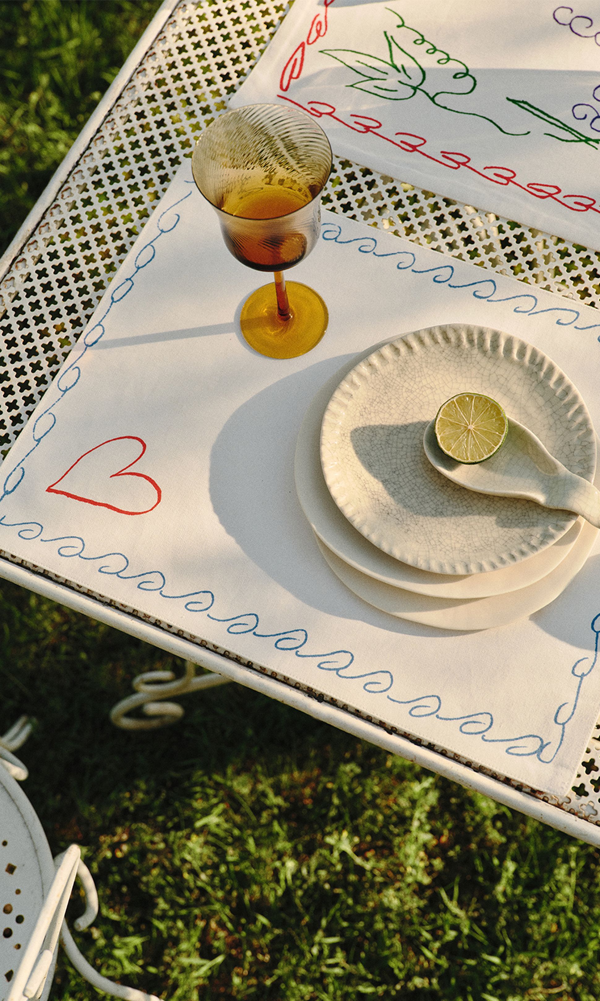 Une table d'extérieur joliment dressée, avec un verre jaune, des assiettes blanches, une rondelle de citron vert et une nappe brodée aux couleurs vives, le tout sur une table en métal.