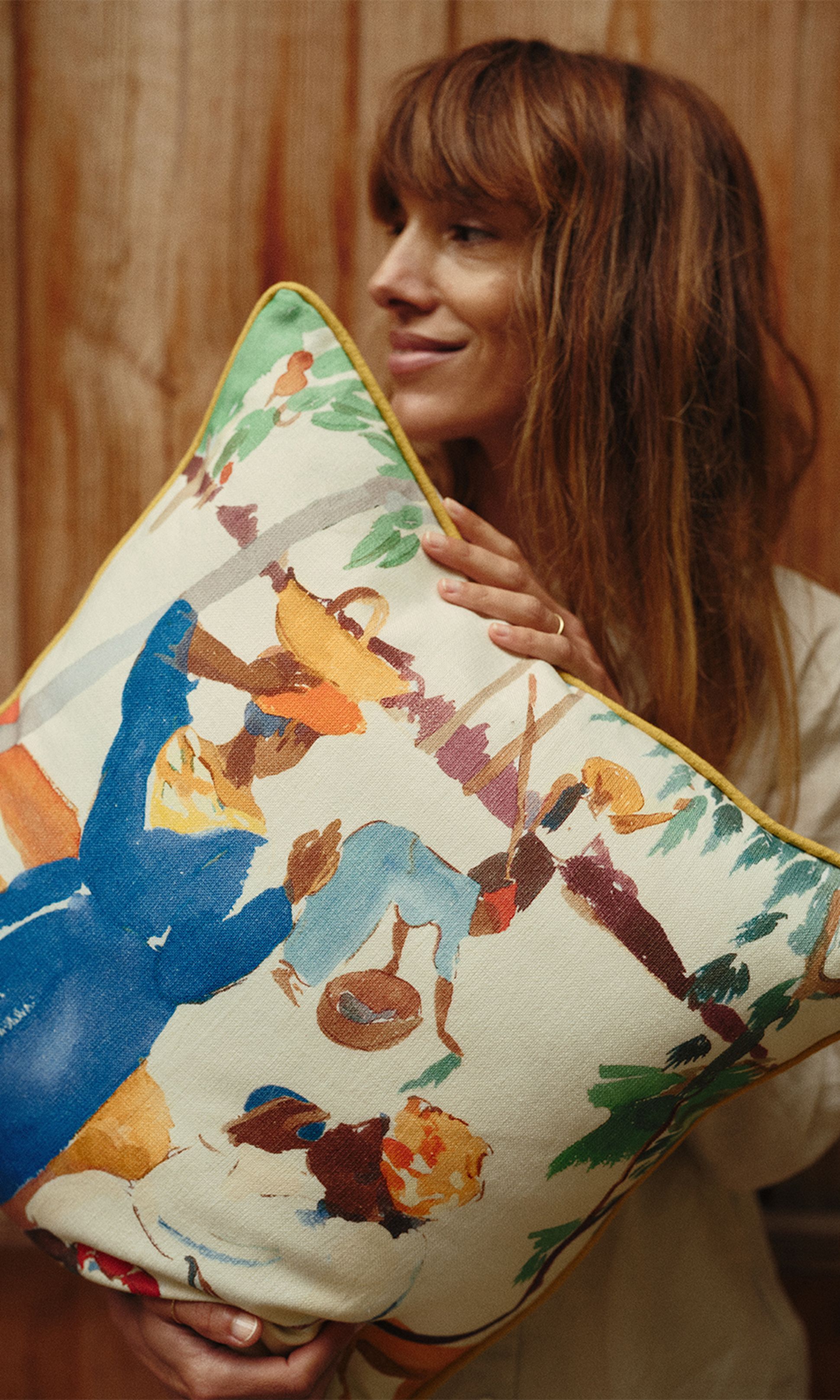 Une femme tenant un coussin coloré orné d'une peinture abstraite et vivante représentant des personnages, sur fond de bois.
