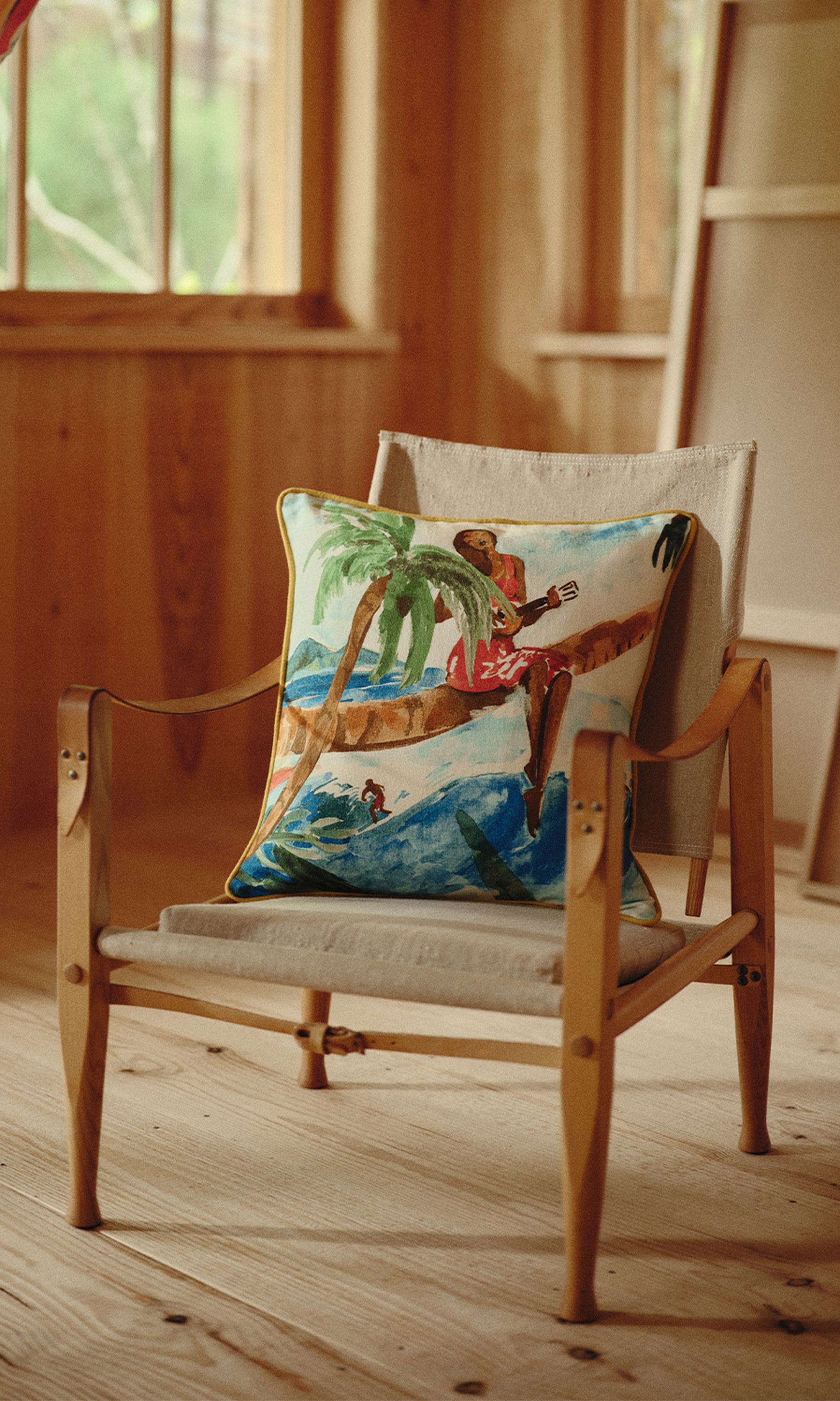 Chaise en bois avec un coussin coloré représentant un paysage tropical, dans une pièce baignée de soleil aux murs et au sol en bois.