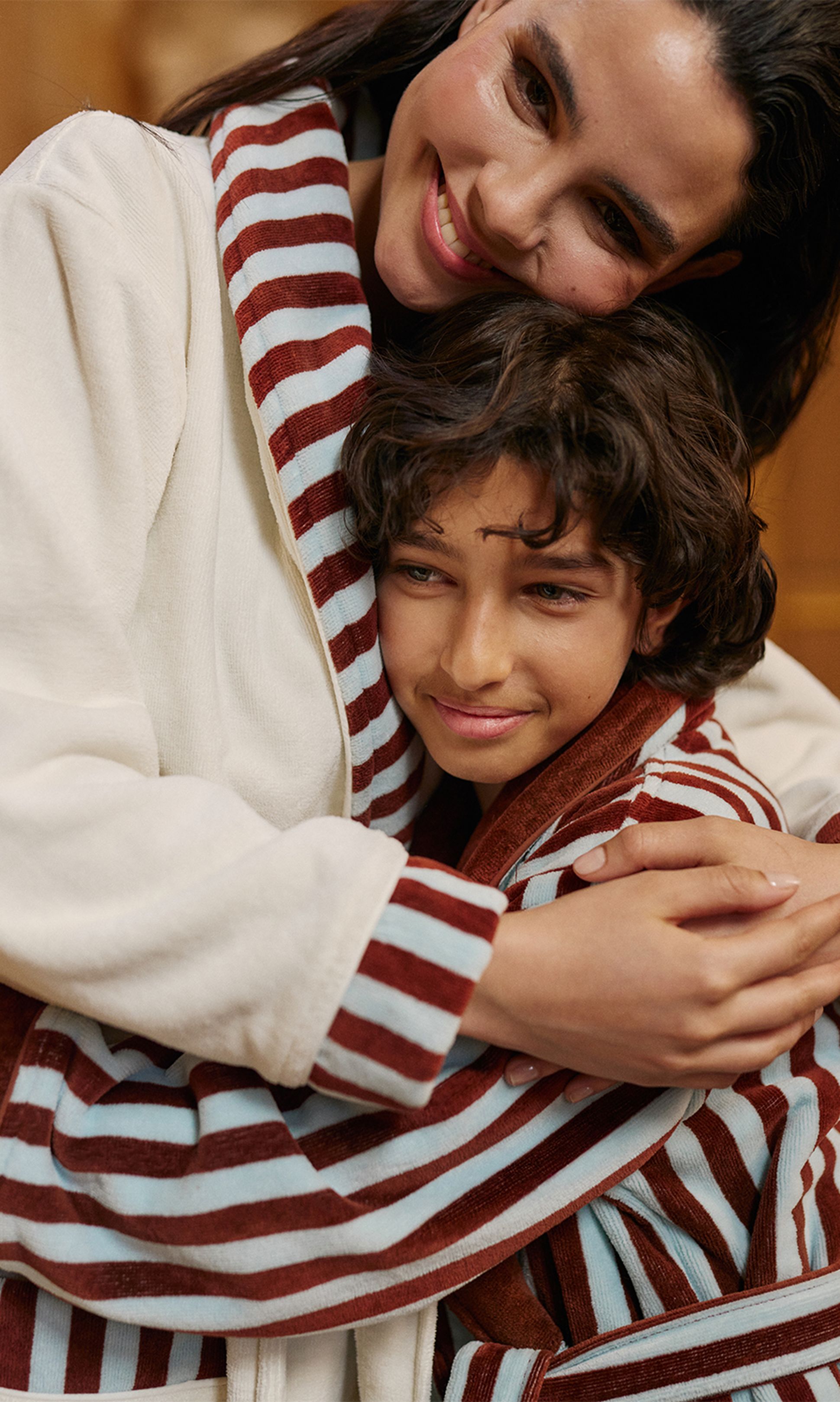 Une femme et un enfant s'étreignent chaleureusement ; tous deux portent des peignoirs rayés de blanc et de rouge brunâtre, et arborent des expressions joyeuses.