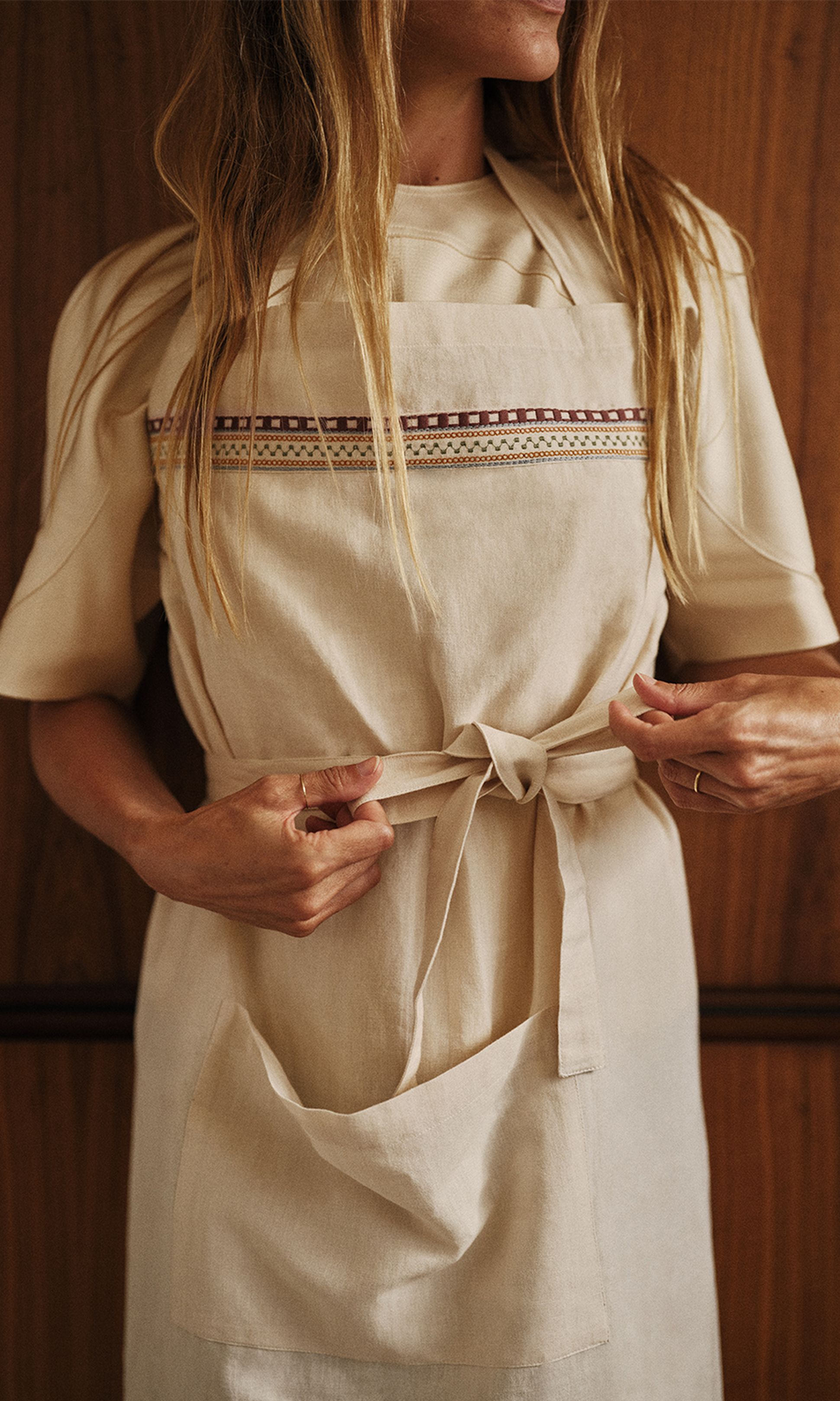 Une femme en train de faire un nœud sur un tablier beige orné de surpiqûres décoratives, debout devant un fond en bois.