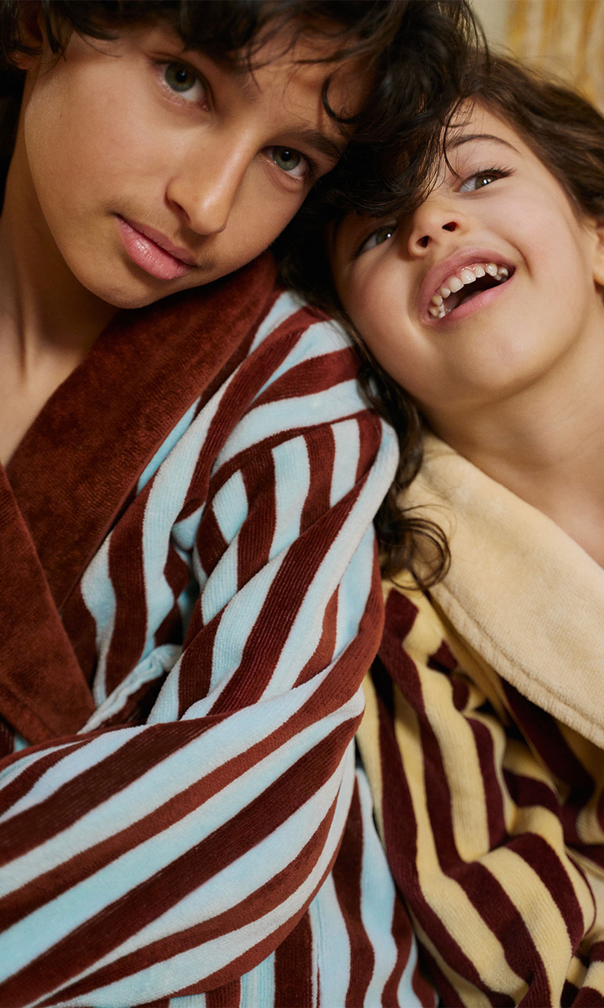 Deux enfants vêtus de peignoirs rayés bien douillets, l'un bleu et marron, l'autre jaune et marron, partagent un moment de joie et de complicité.