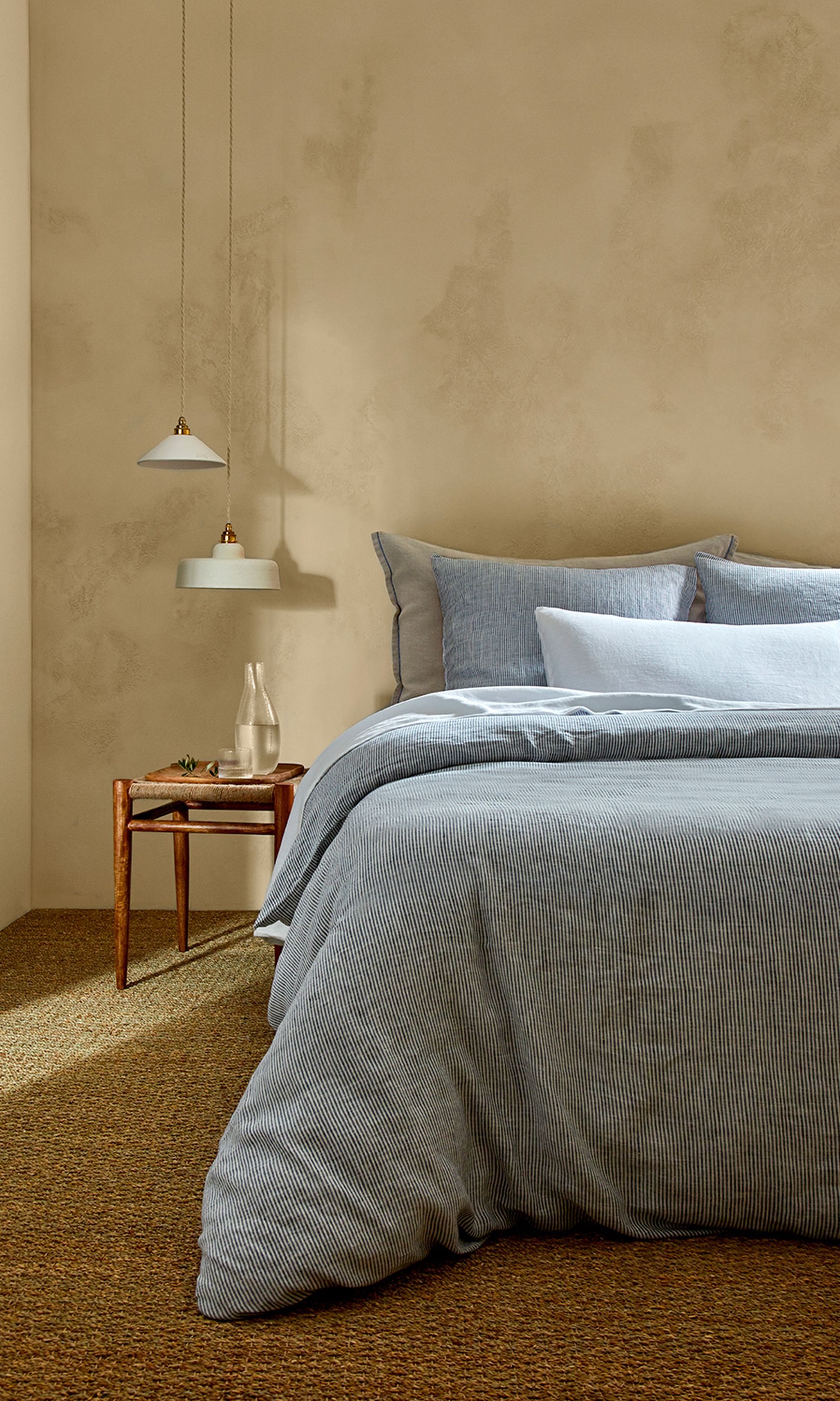 Chambre à coucher chaleureuse, avec une couette bleue à rayures, des oreillers et une table de chevet en bois sur laquelle repose une carafe en verre. Des suspensions modernes sont accrochées au plafond.