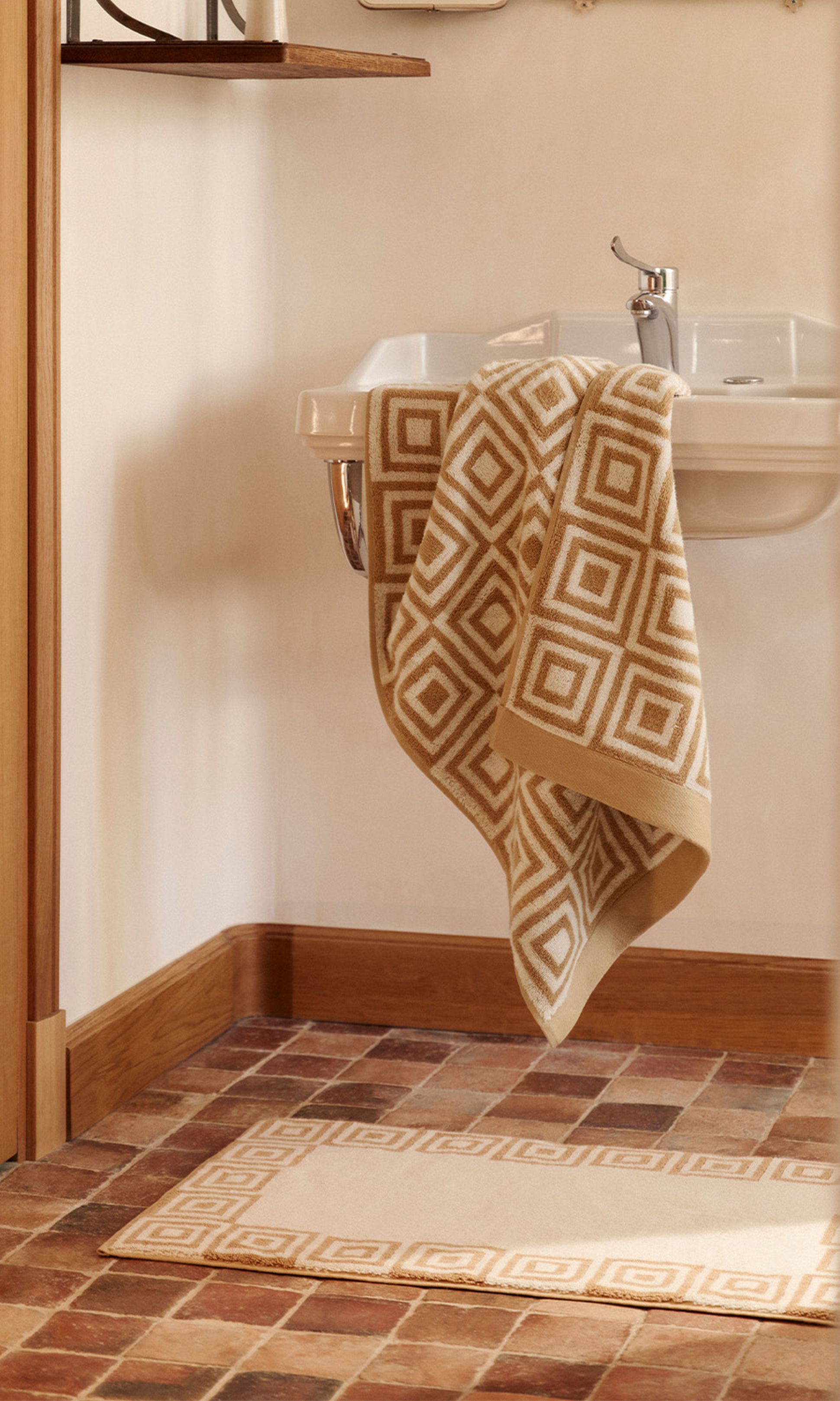 Salle de bain avec une serviette à motifs géométriques suspendue à un lavabo blanc. Tapis assorti sur un sol en carreaux de terre cuite. Murs aux tons neutres et accents en bois.