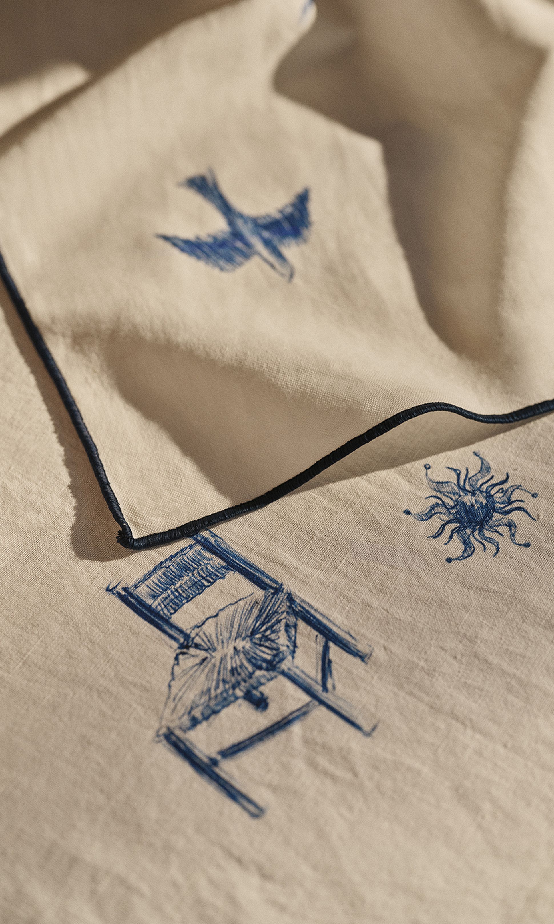 Gros plan sur un tissu beige orné de motifs brodés bleus représentant un oiseau, une forme ressemblant à un soleil et une chaise, le tout bordé d'une passementerie foncée.