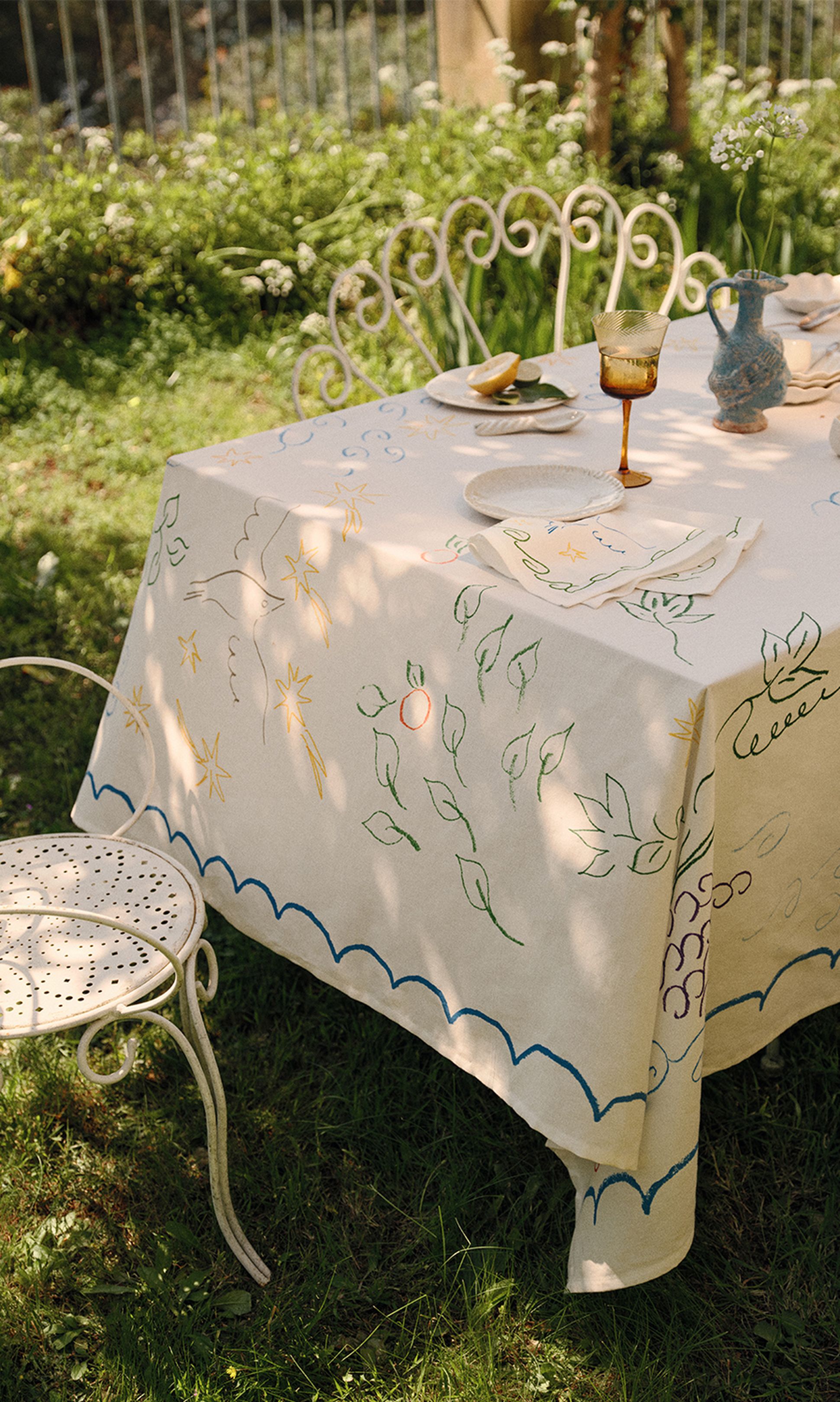 Une table de pique-nique recouverte d'une nappe brodée aux couleurs vives, avec des assiettes, une boisson et un vase de fleurs, dans un jardin ensoleillé.