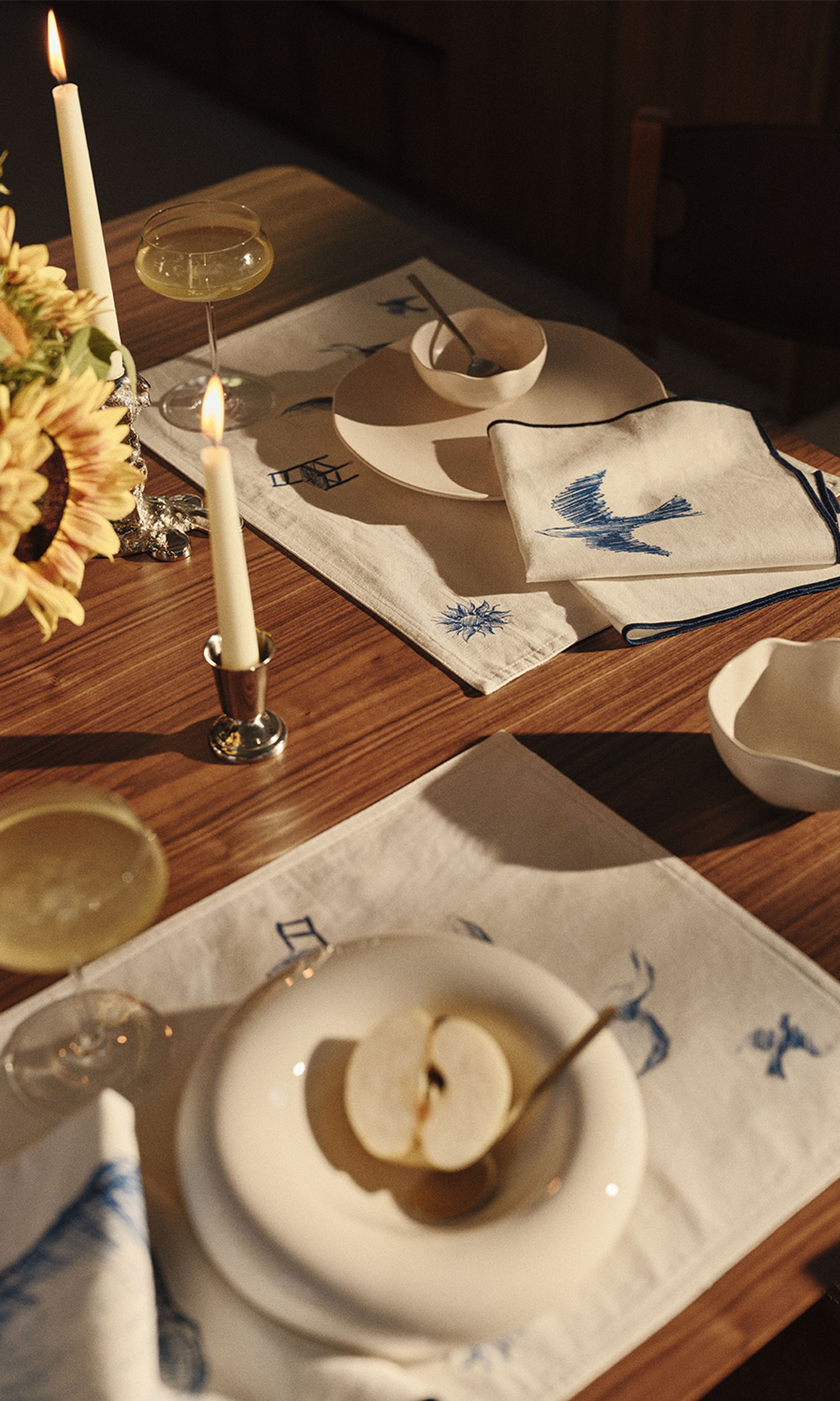 Une table en bois dressée avec des assiettes blanches, une pomme coupée en deux, des coupes à champagne, des serviettes brodées, des tournesols et une bougie allumée.