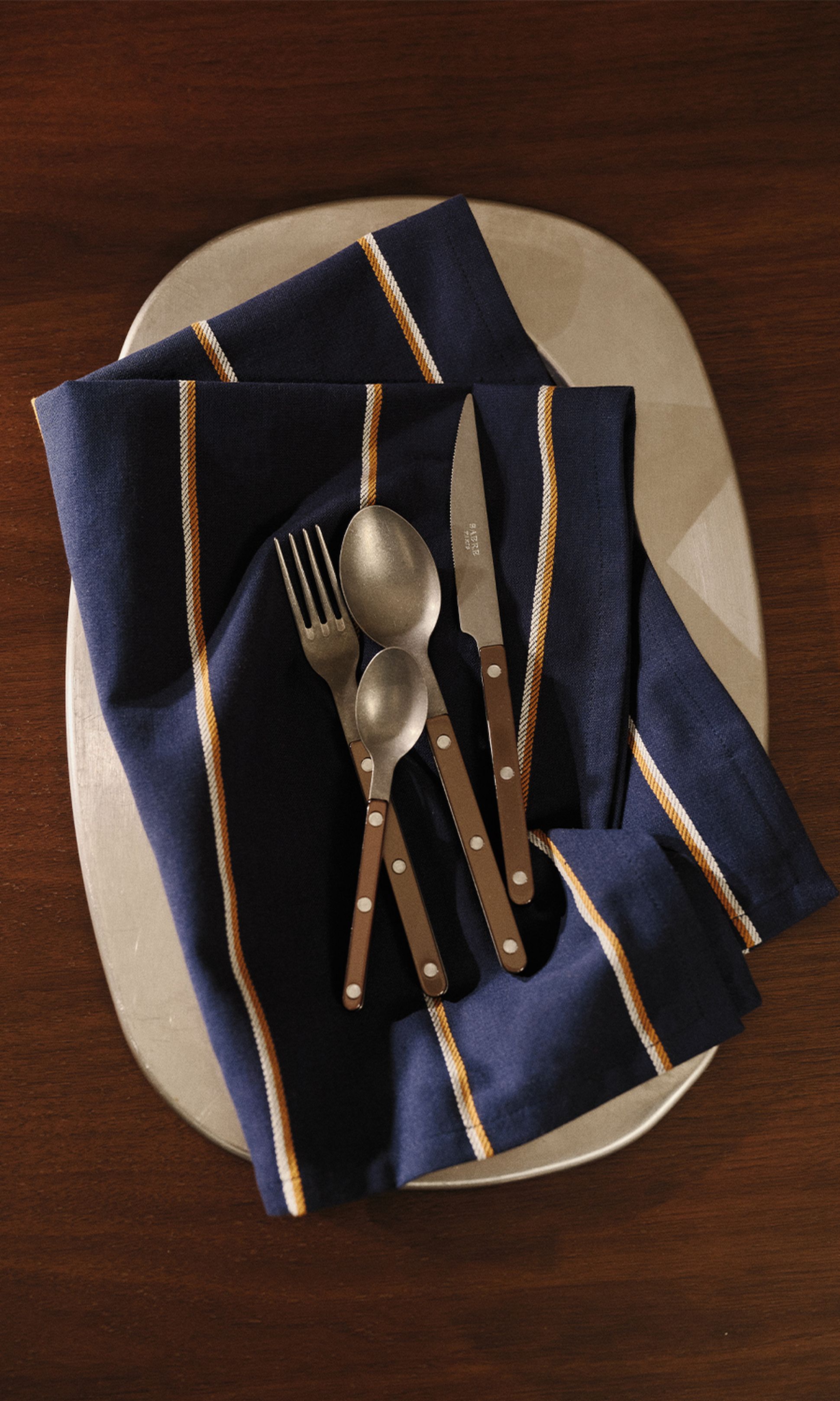 Un service de couverts posé sur une serviette bleue rayée, sur une assiette, le tout disposé sur une table en bois.