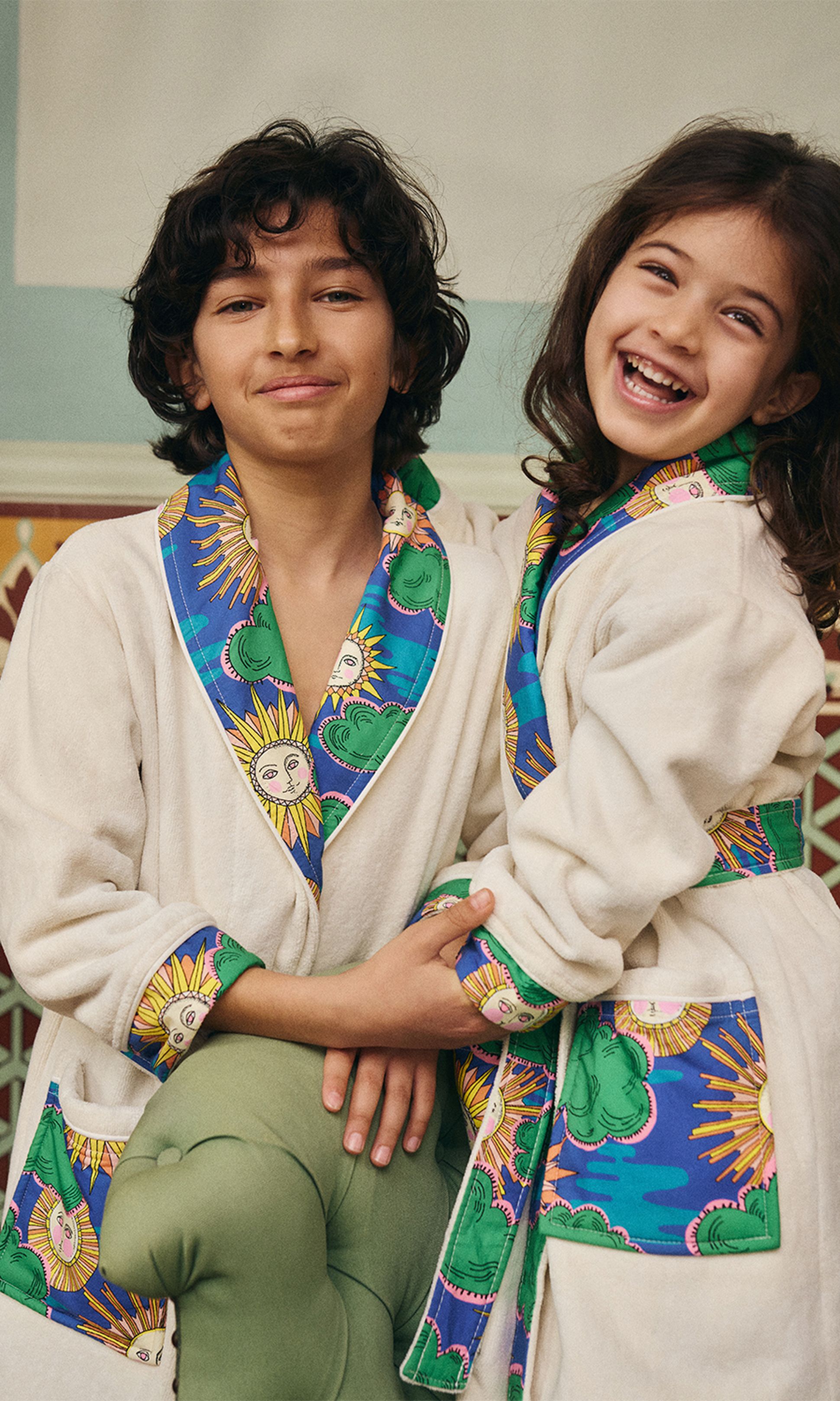 Deux enfants vêtus de robes assorties ornées de motifs solaires colorés sourient joyeusement, enlacés dans un décor chaleureux.