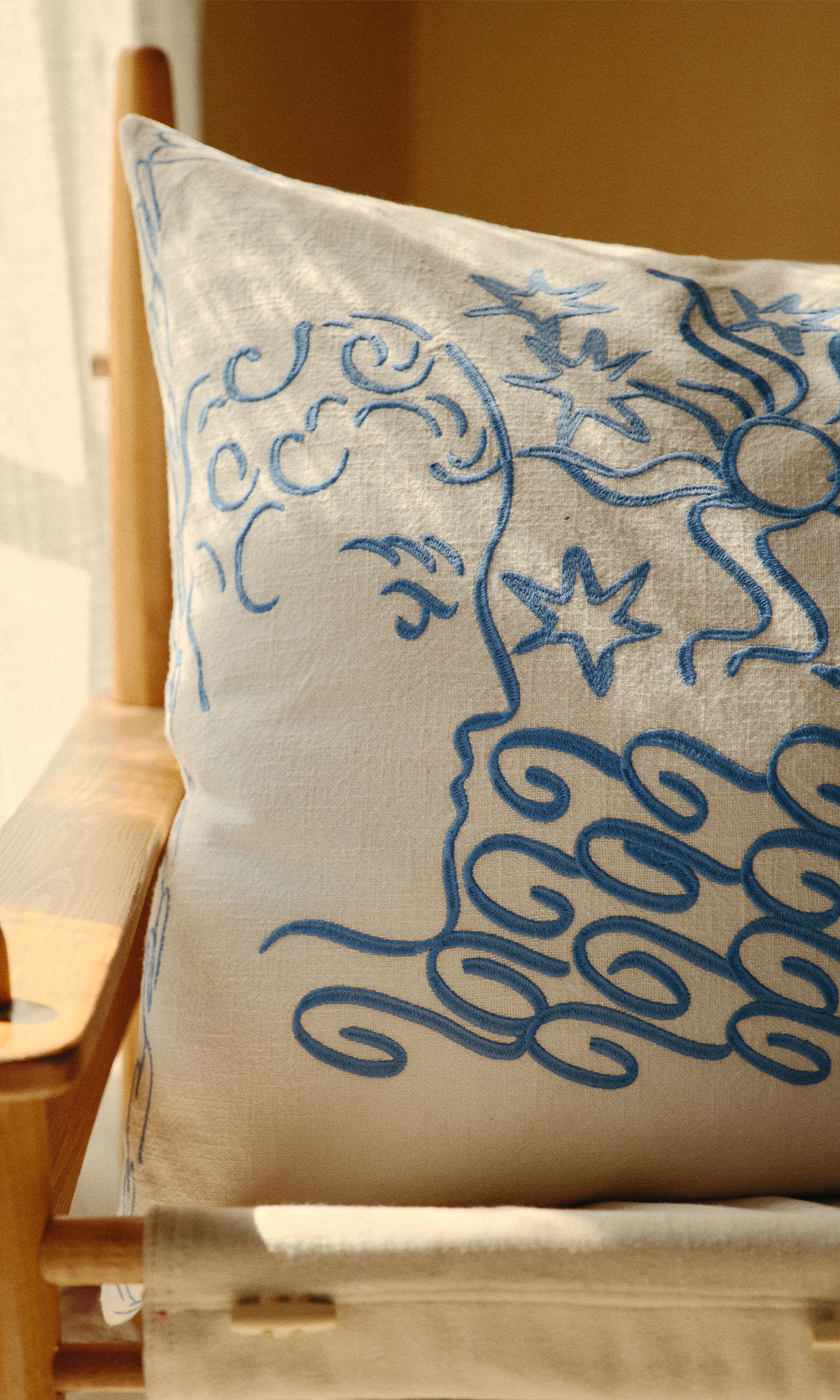 Un coussin blanc orné d'un visage abstrait et de volutes brodés en bleu, posé sur une chaise en bois, baigné par la lumière naturelle du soleil.