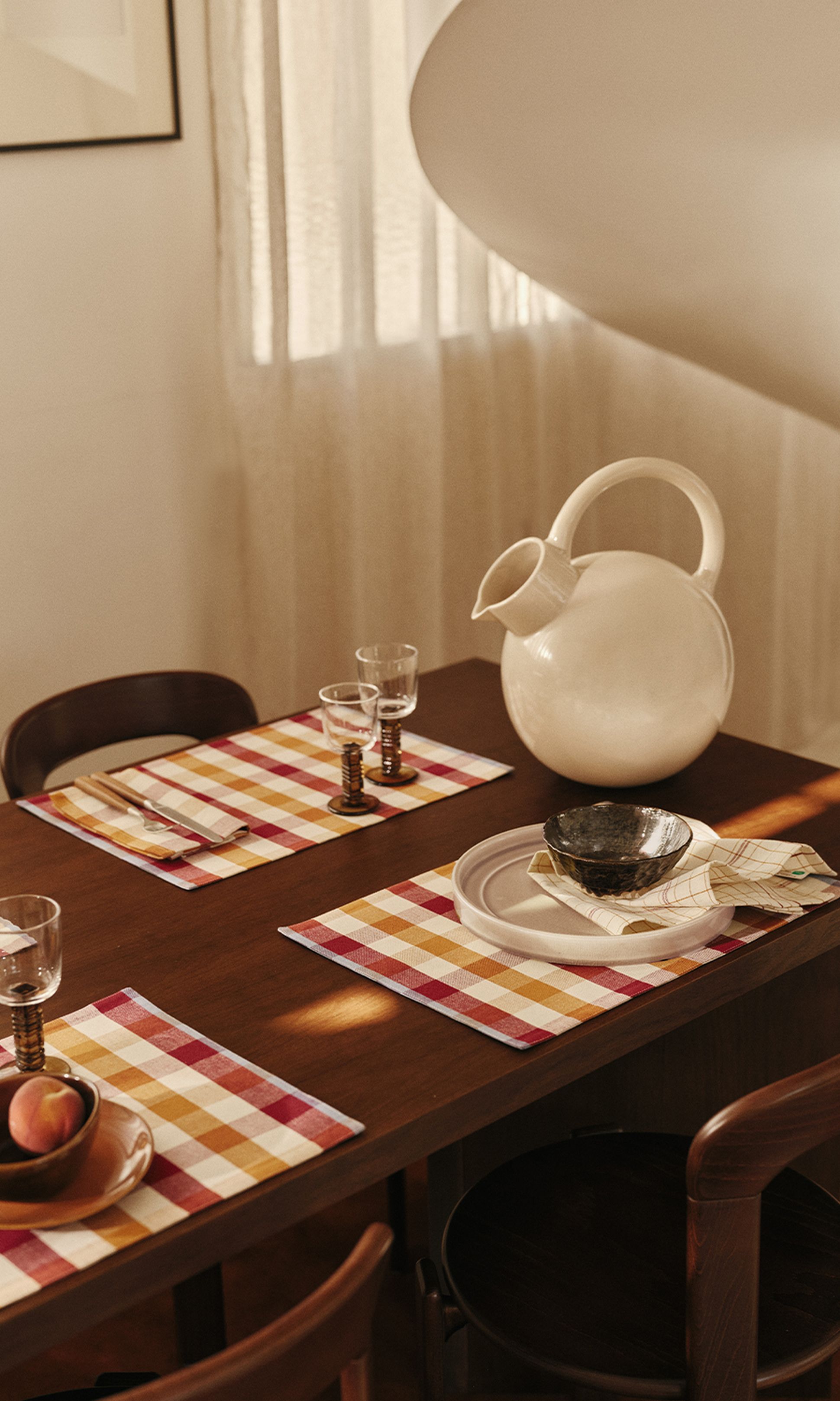 Une table à manger dressée avec des sets de table à carreaux, des verres, un pichet rond blanc et des bols, baignée d'une douce lumière naturelle filtrant à travers des rideaux vaporeux.